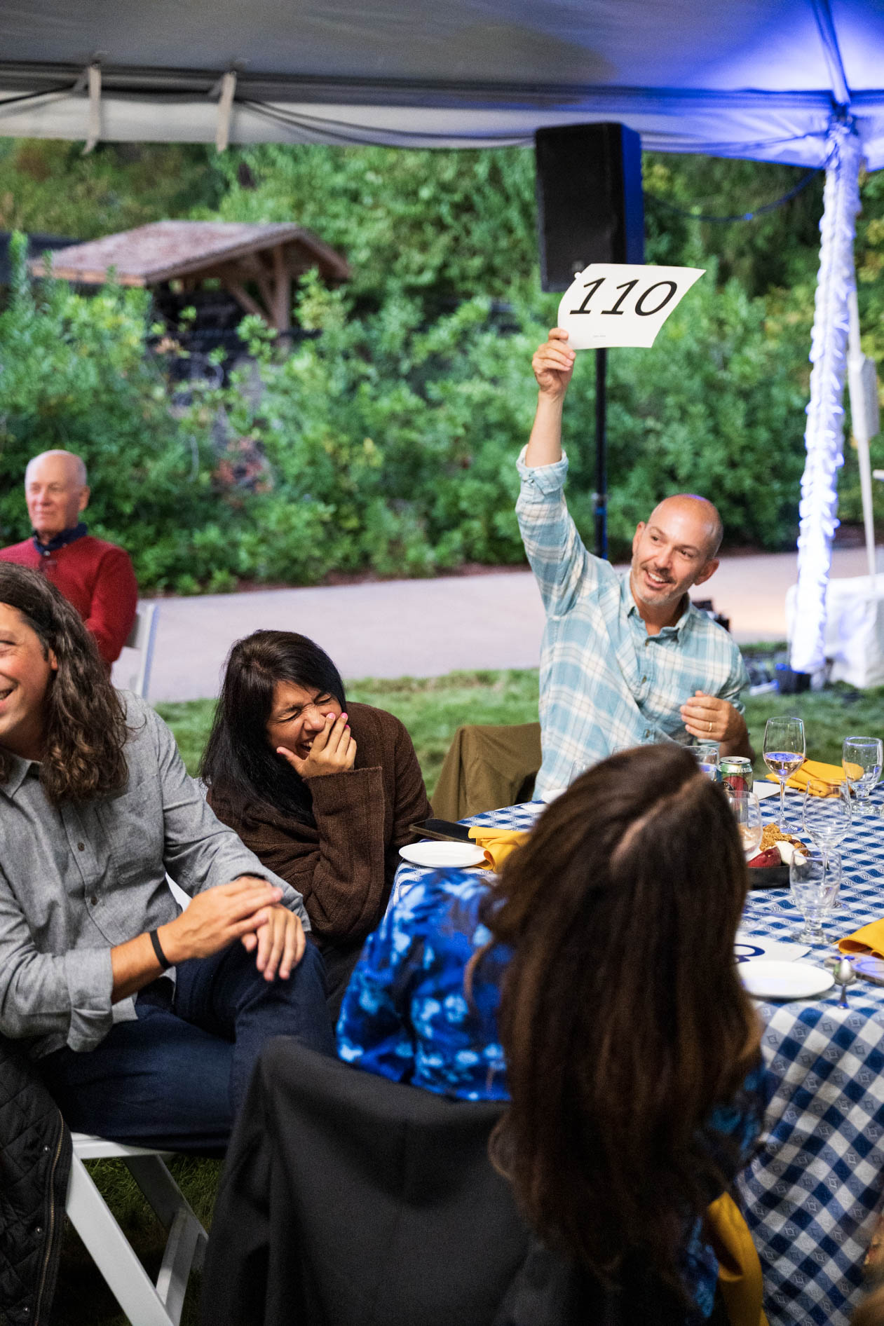 A lively group of guests enjoys an auction at the Feast in the Garden event in SE Portland. A man at the table holds up a number card, smiling and participating in the fundraiser, as other guests laugh and share a moment of joy.