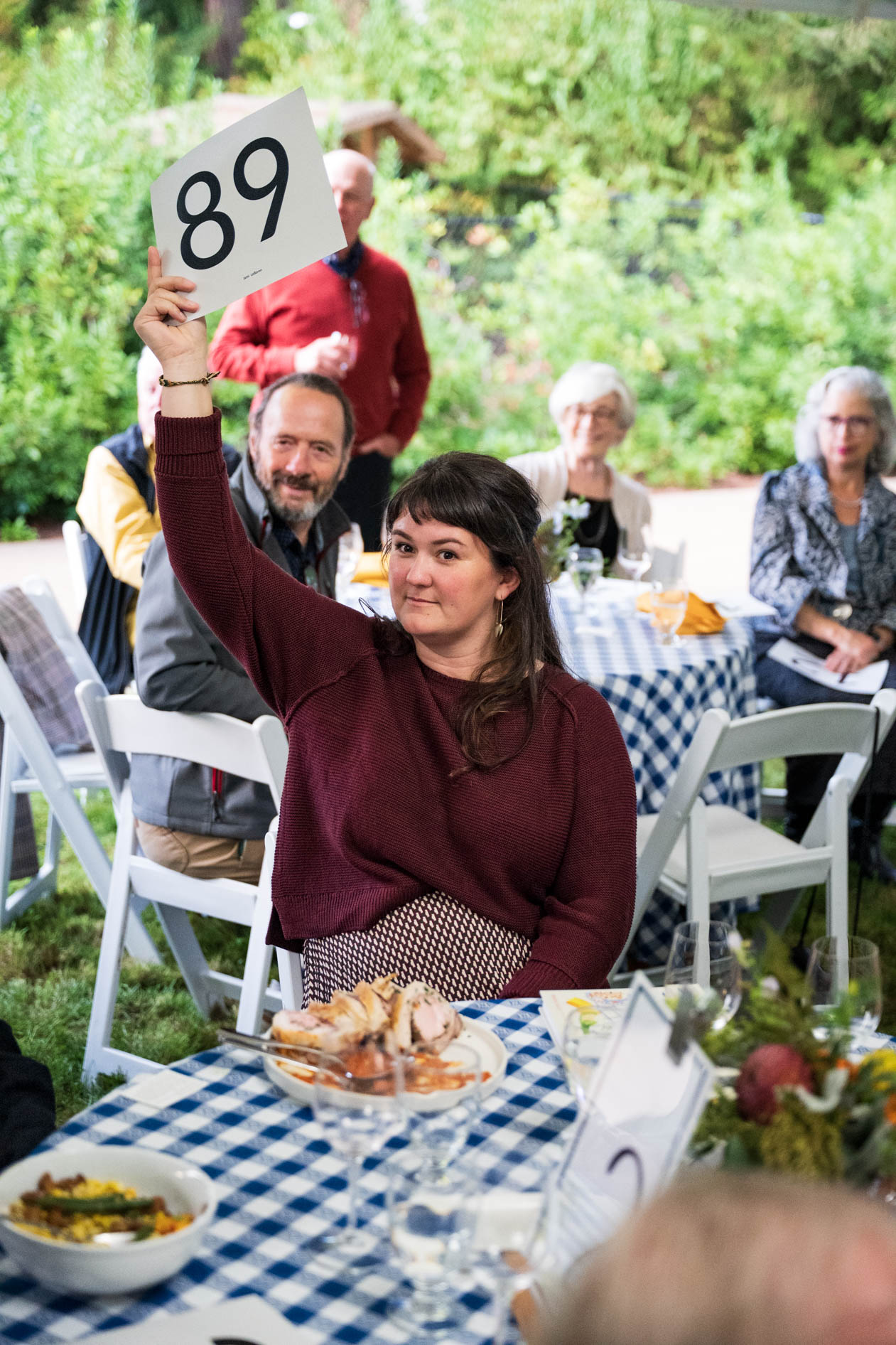 A woman holds up an auction number card, participating in the lively fundraiser at the Feast in the Garden event in SE Portland. She looks at the camera with a confident smile, surrounded by other guests enjoying the meal.