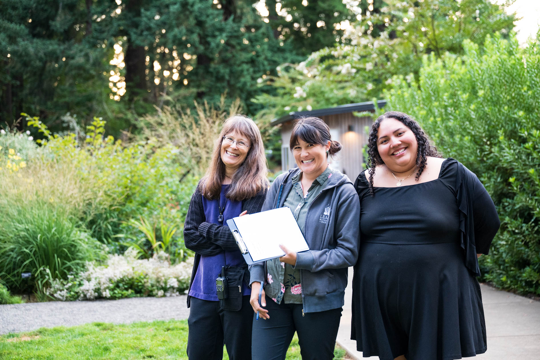 A smiling group of three women, two of whom appear to be event staff, pose for a photo. They are standing on a lush lawn, holding a clipboard, capturing a warm and collaborative moment at the Feast in the Garden event in SE Portland.