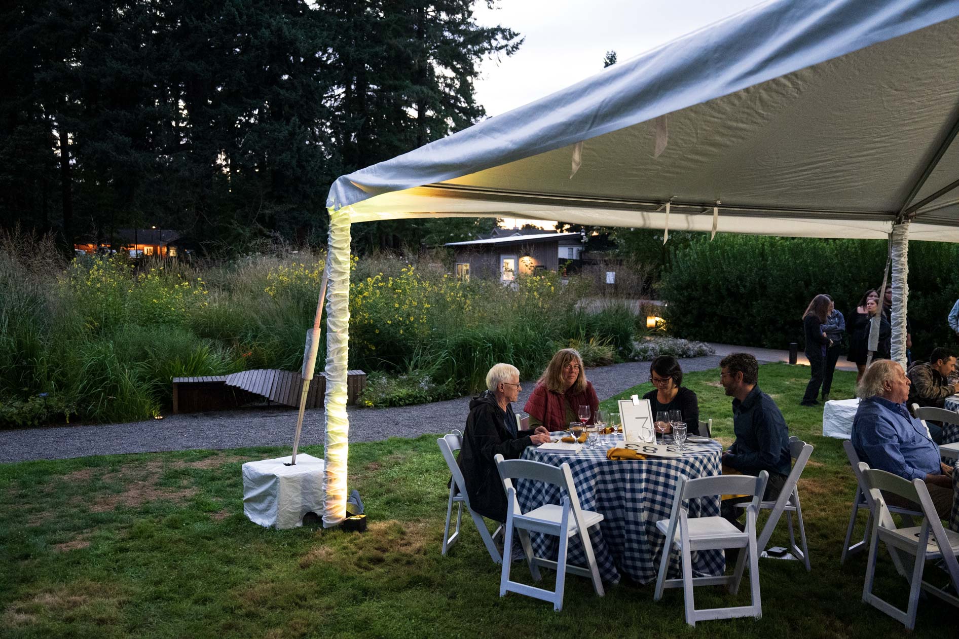 A peaceful table of four guests is seated under a white tent on a lawn, enjoying drinks and conversation. The scene captures an intimate moment at the Feast in the Garden event in SE Portland at dusk, with ambient light from the garden pathways.