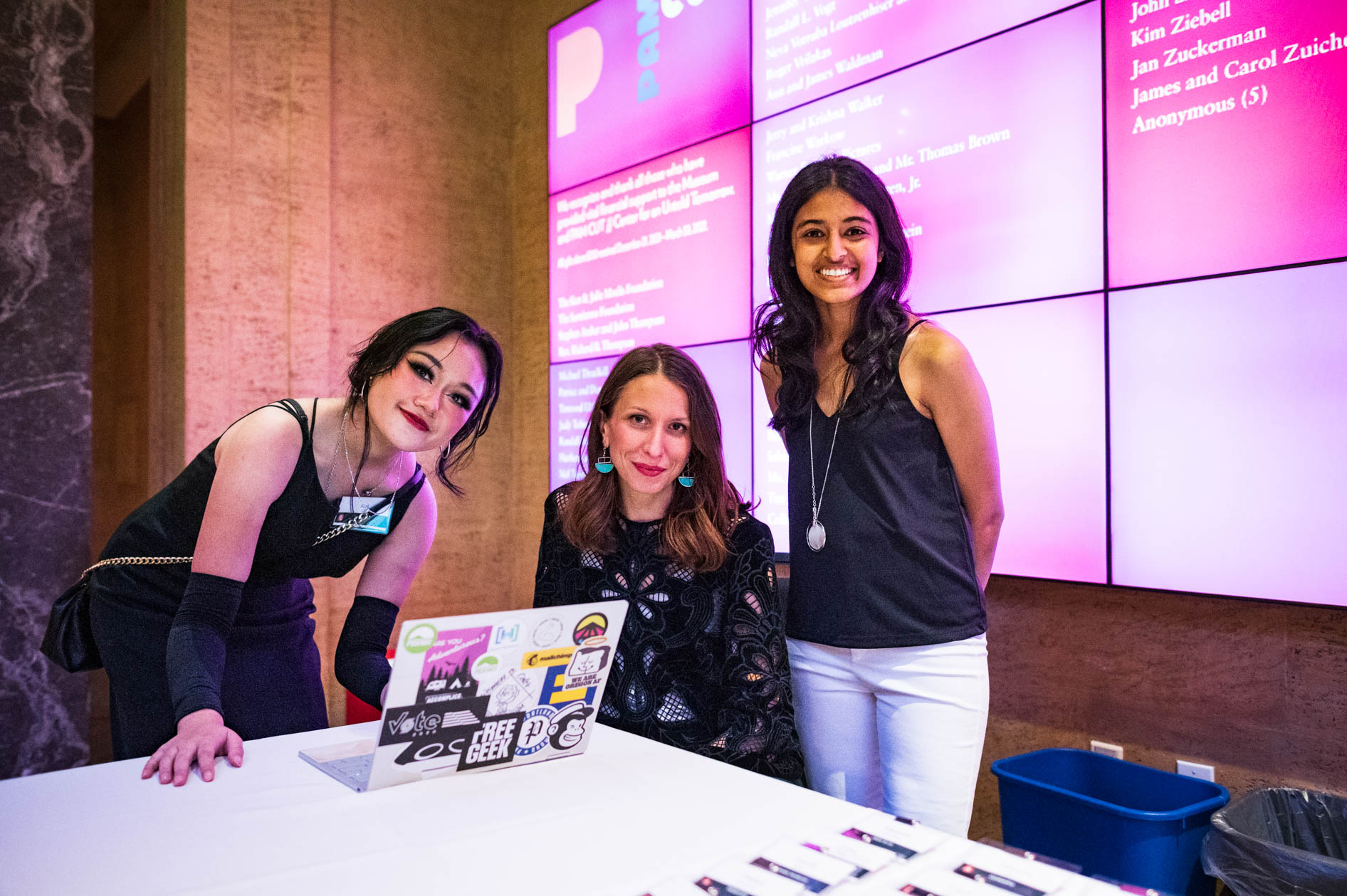 Three women smile from a check-in table, with a large screen displaying the OEN Awards logo behind them, capturing a moment of welcome at the Portland Art Museum.