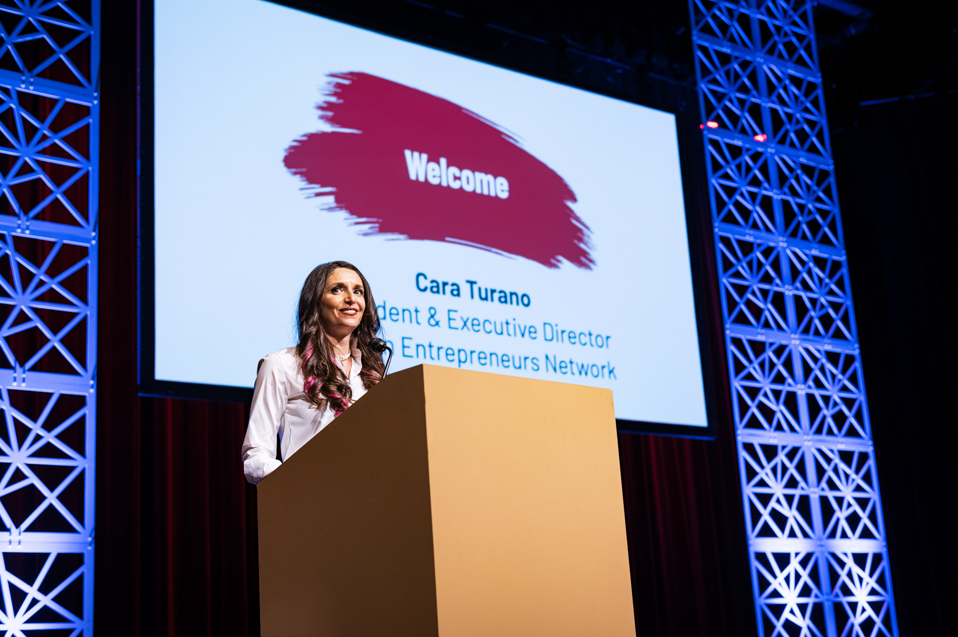 Cara Turano, President & Executive Director of the Oregon Entrepreneurs Network, smiles at the audience from a stage. She is welcoming guests to the OEN Awards in Portland with her name on the screen behind her.