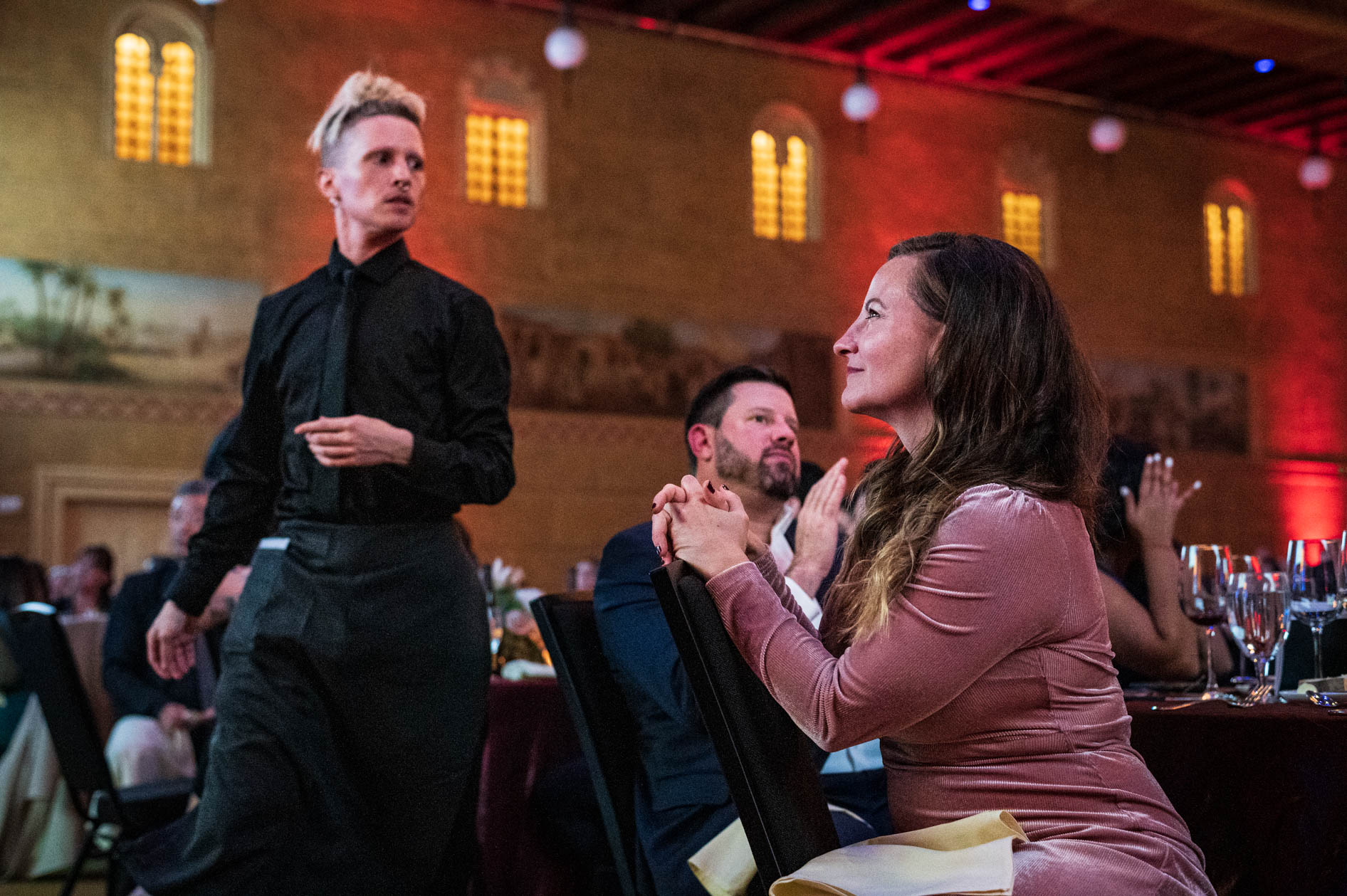 A stylish waitress with a blonde hairstyle is serving guests at a table. A woman in a pink dress looks up with a smile as she is served, while other attendees clap in the background, capturing a moment of warm service at the OEN Awards in Portland.