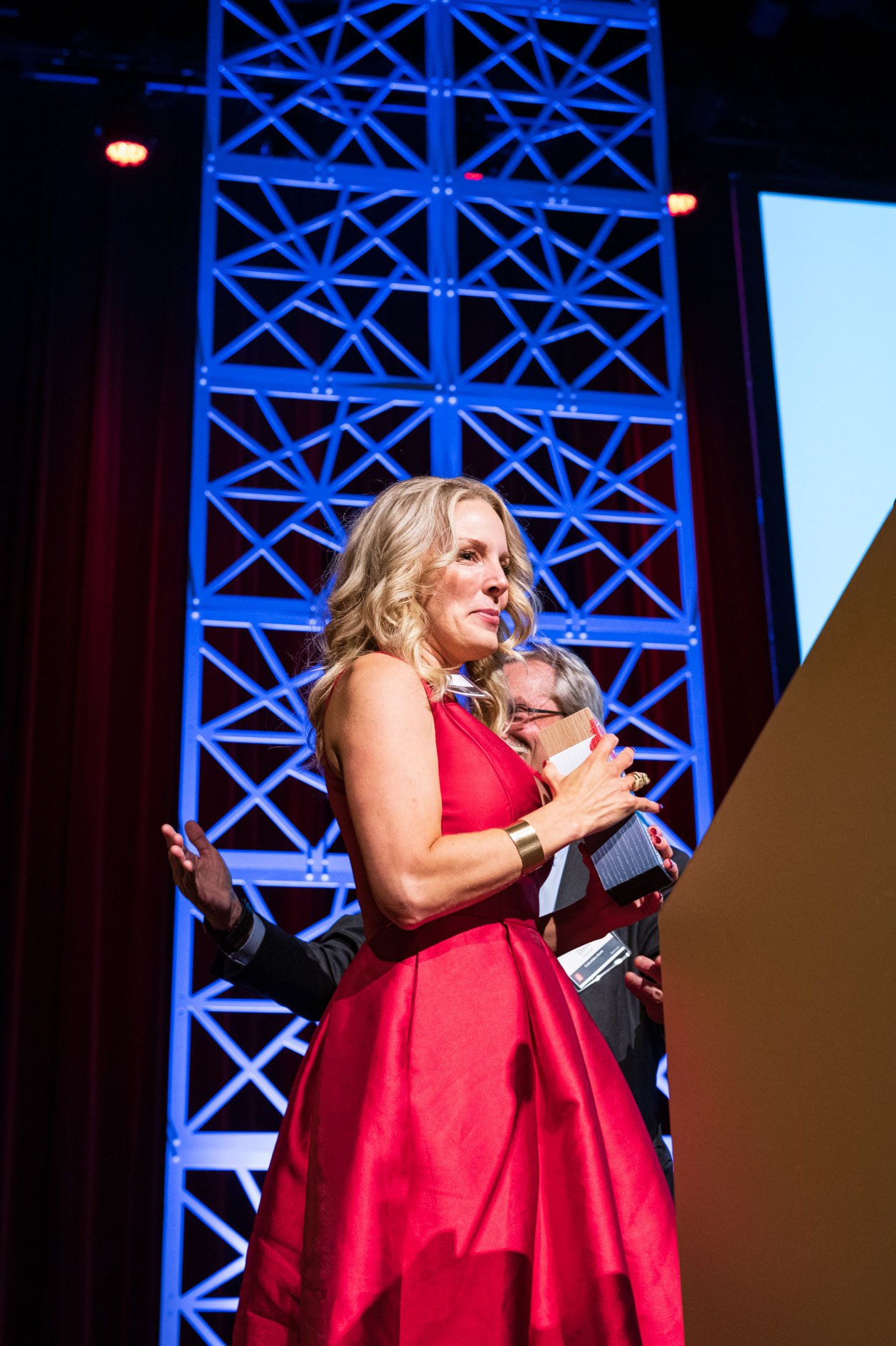 A woman in a vibrant red dress holds an award on stage. She smiles with a triumphant expression, capturing a moment of success and celebration at the OEN Entrepreneurship Awards in downtown Portland.