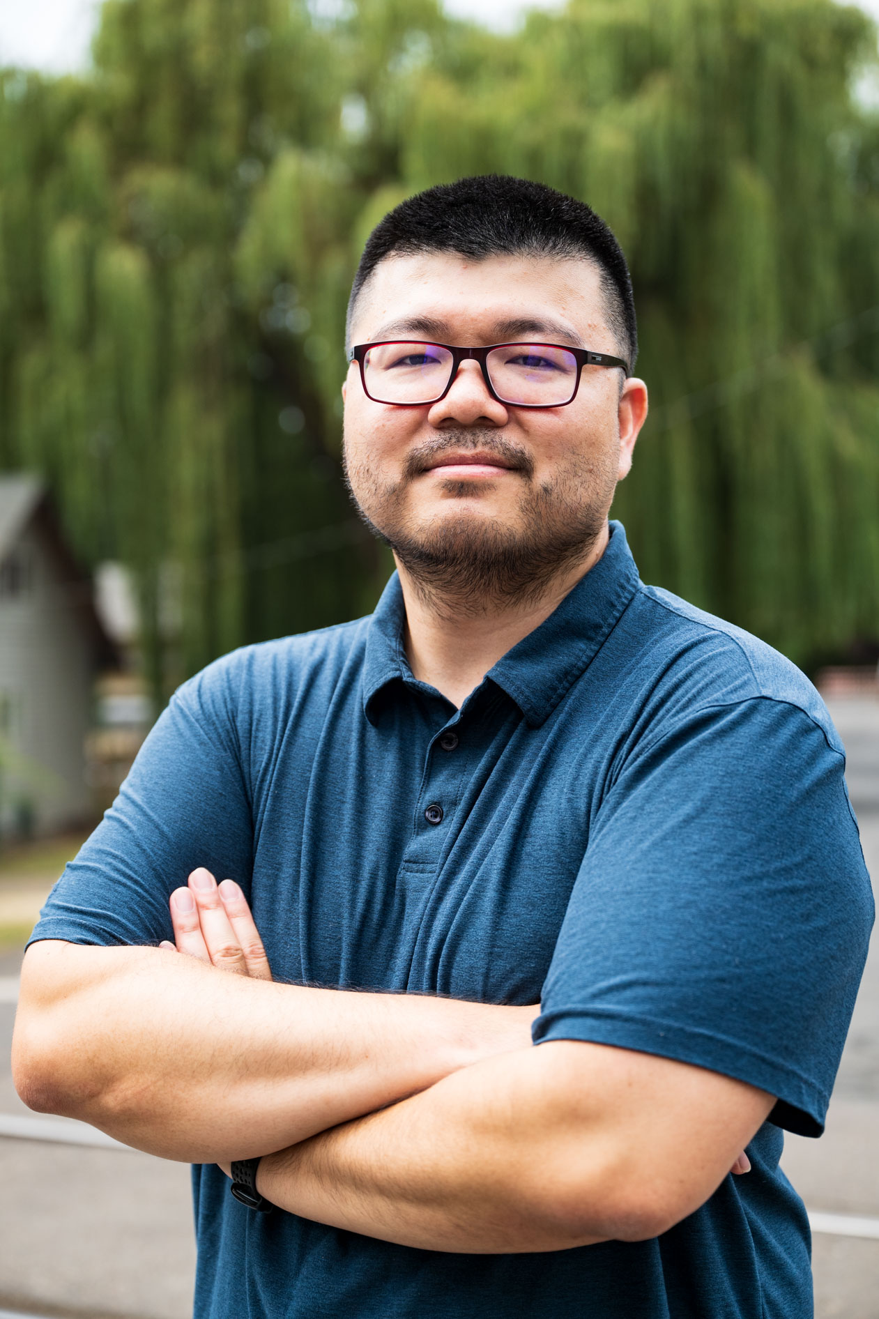 A professional and authentic portrait of a smiling person with glasses and a blue shirt, posing with their arms crossed. This is an example of commercial photography for a business coaching company in Portland, Oregon.
