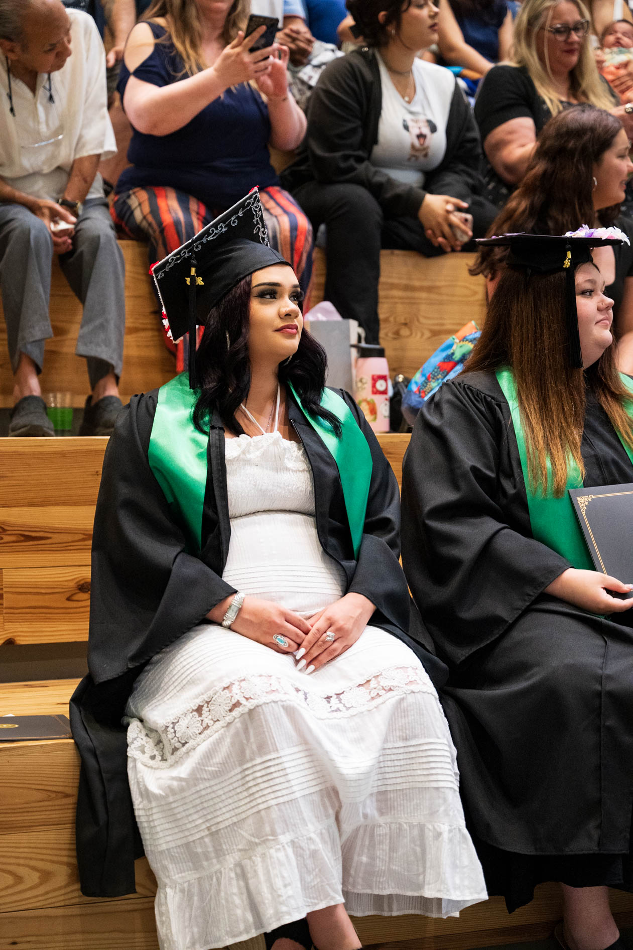A candid, documentary-style photograph of a graduate in a cap and gown sitting on bleachers during a graduation ceremony. This is an example of event photography for Open School East in Portland, Oregon.