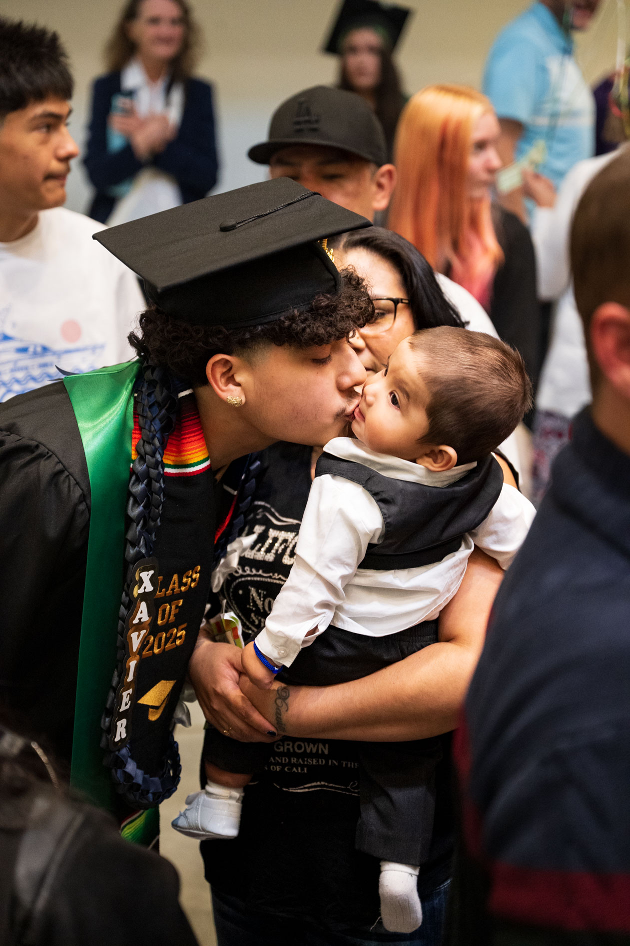 A graduate in a cap and gown kisses a baby on the cheek at a graduation ceremony. This is an example of an event photographer in Portland, Oregon, capturing a joyful, multi-generational moment for Open School East.