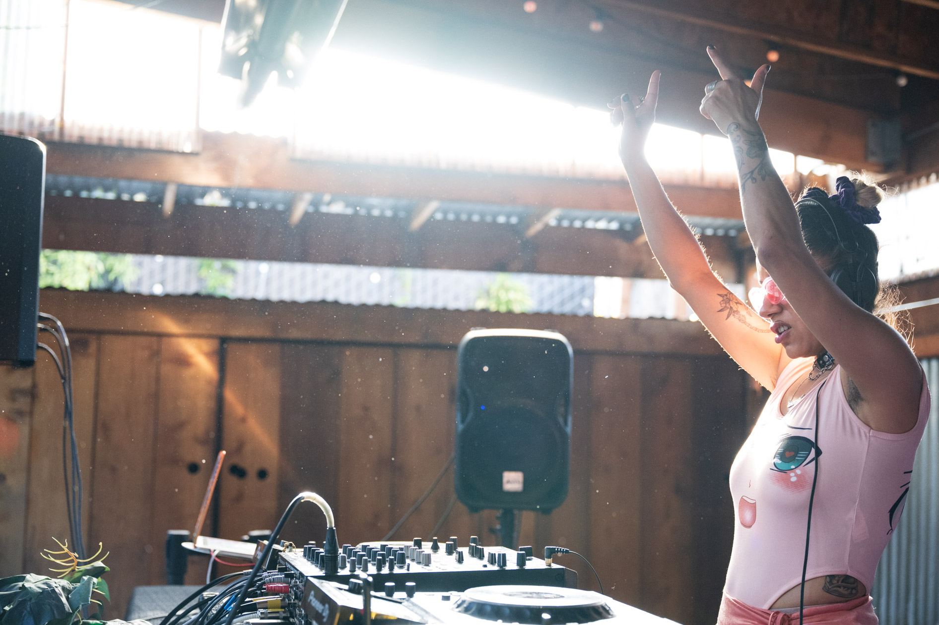 A candid photo of DJ Lapaushi performing a set at a live outdoor event. The DJ is wearing pink sunglasses and has their arms raised in the air. This is an example of commercial photography for an artist's portfolio.