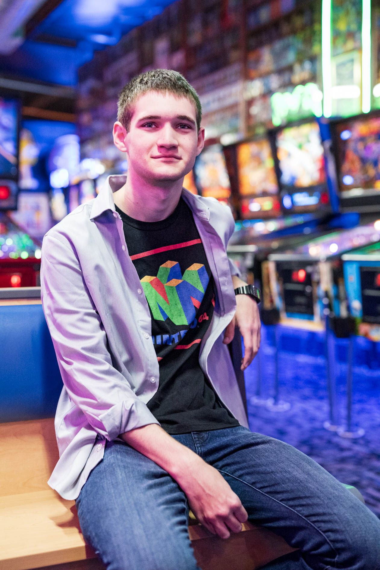 A relaxed senior portrait of a person in a Nintendo 64 shirt, sitting in a brightly lit arcade in Hillsboro, Oregon.
