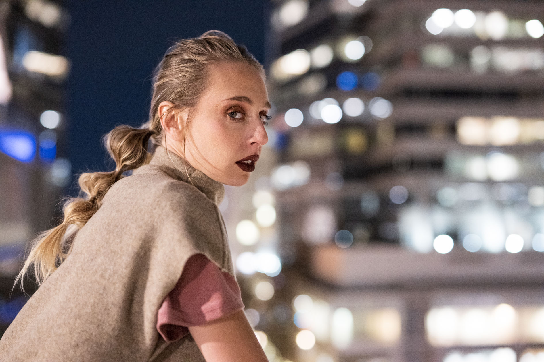 A commercial portrait of a female model with a braided ponytail and a poncho from the boutique brand Ale O, posing at night with the city lights in the background. This image showcases lifestyle brand photography in Portland, Oregon.
