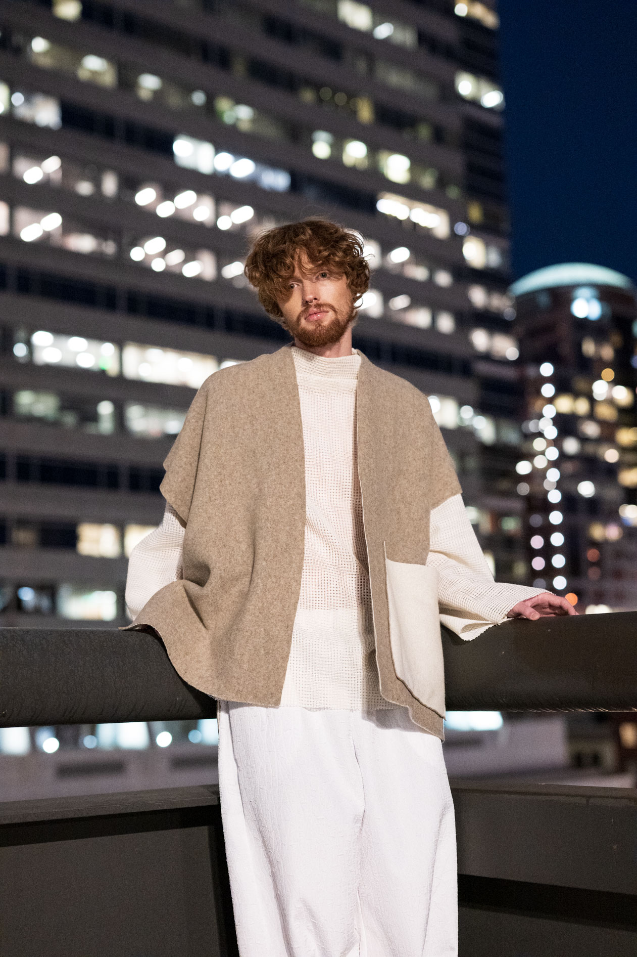 A commercial portrait of a male model with reddish hair and beard, wearing a beige poncho over a white knit shirt and pants from the boutique brand Ale O, posing at night with the city lights in the background.