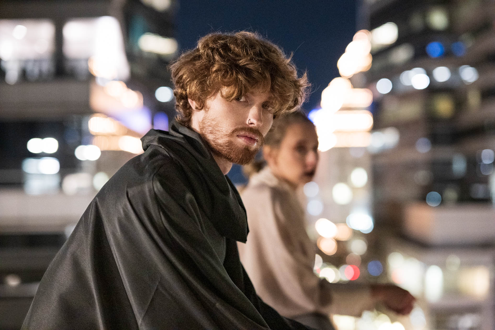 A commercial portrait of a male model with reddish hair and a beard, wearing a black poncho from the boutique brand Ale O, posing at night with the city lights in the background. This image showcases lifestyle brand photography in Portland, Oregon.