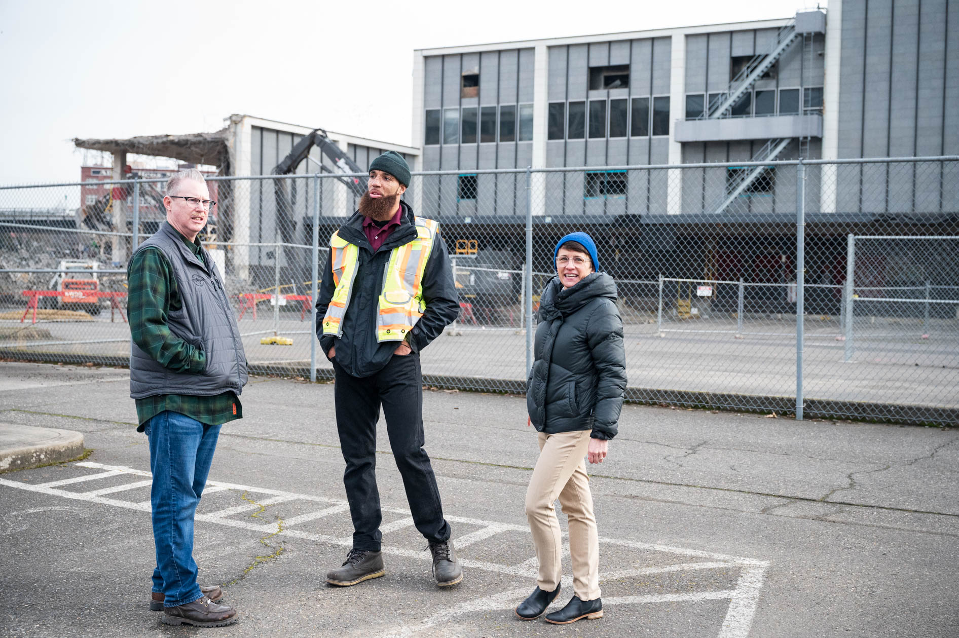 A candid photograph of three attendees talking outside the fenced-off former downtown post office demolition site in Portland, Oregon.