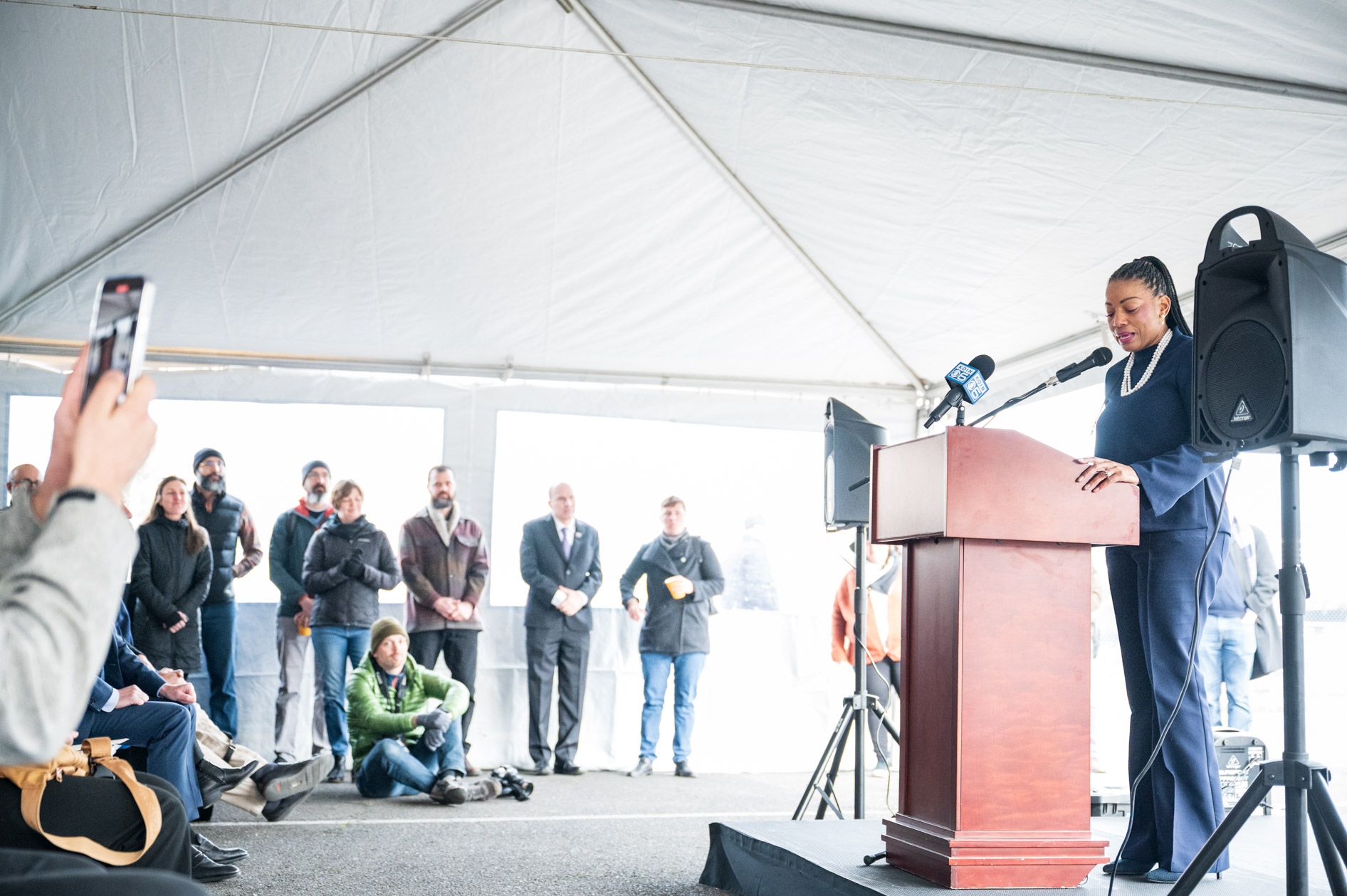 A woman giving a speech at a podium under a white tent to a crowd of attendees at the Prosper Portland demolition event.