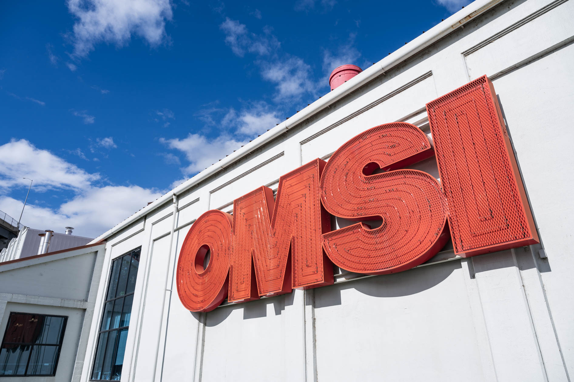 A large red OMSI sign on the white exterior of the building, under a bright blue sky. This showcases the Sneaker Week event venue in Portland, Oregon.