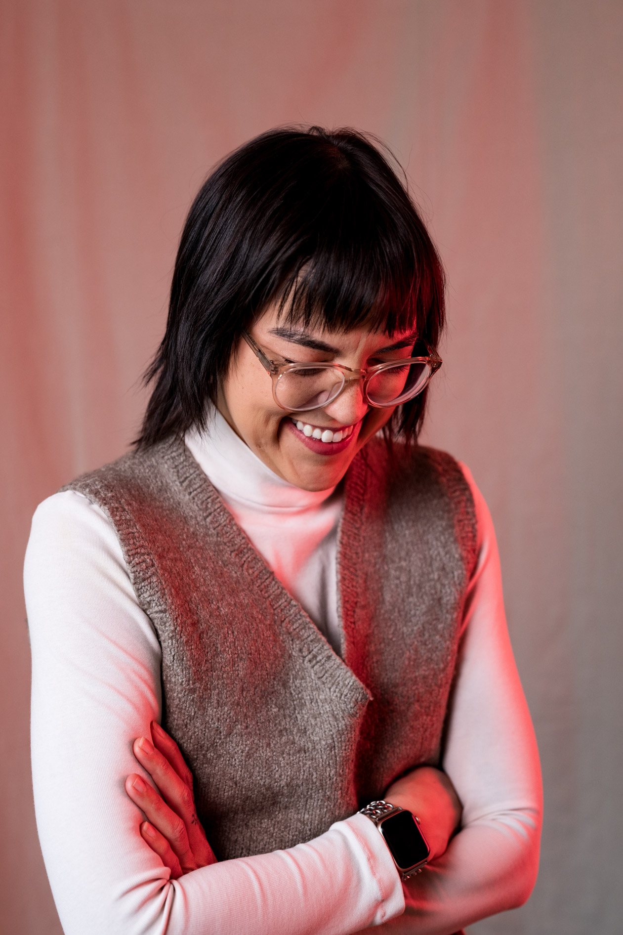 A smiling, candid inclusive portrait of Lee DeJonge with their head down, wearing a knit vest and glasses. This showcases creative portraits in Portland, Oregon.