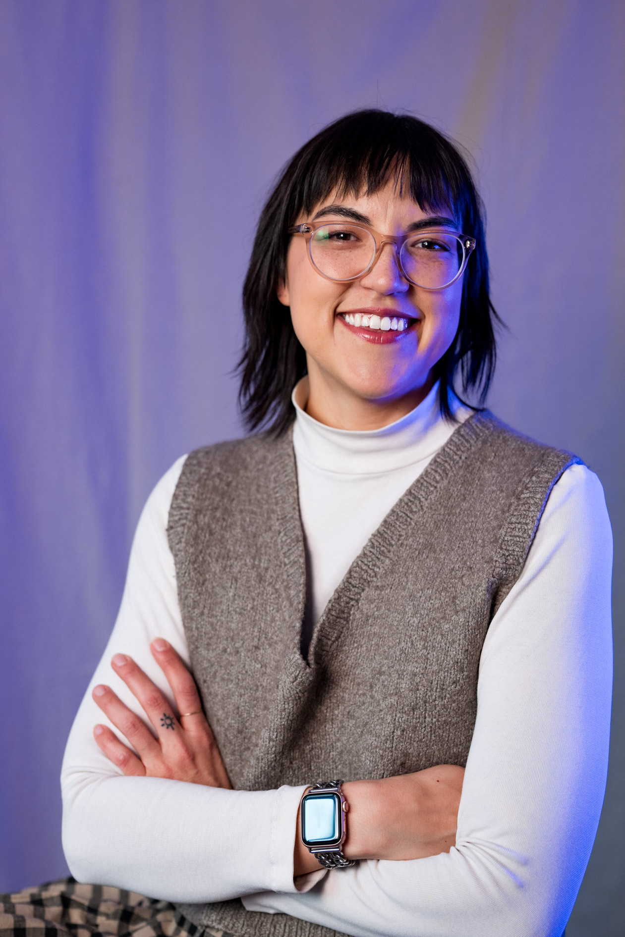 A smiling inclusive portrait of Lee DeJonge with their arms crossed, wearing a knit vest and glasses. This showcases creative portraits in Portland, Oregon.