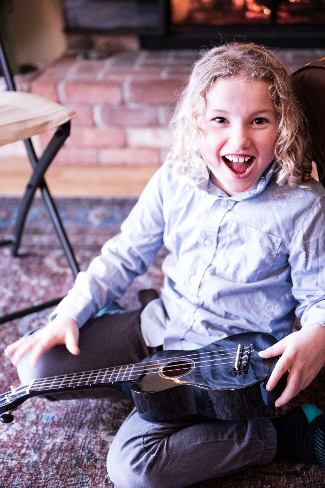 A joyful inclusive family portrait of a child with curly hair and a ukulele, sitting in front of a fireplace during a holiday session in Beaverton, Oregon.