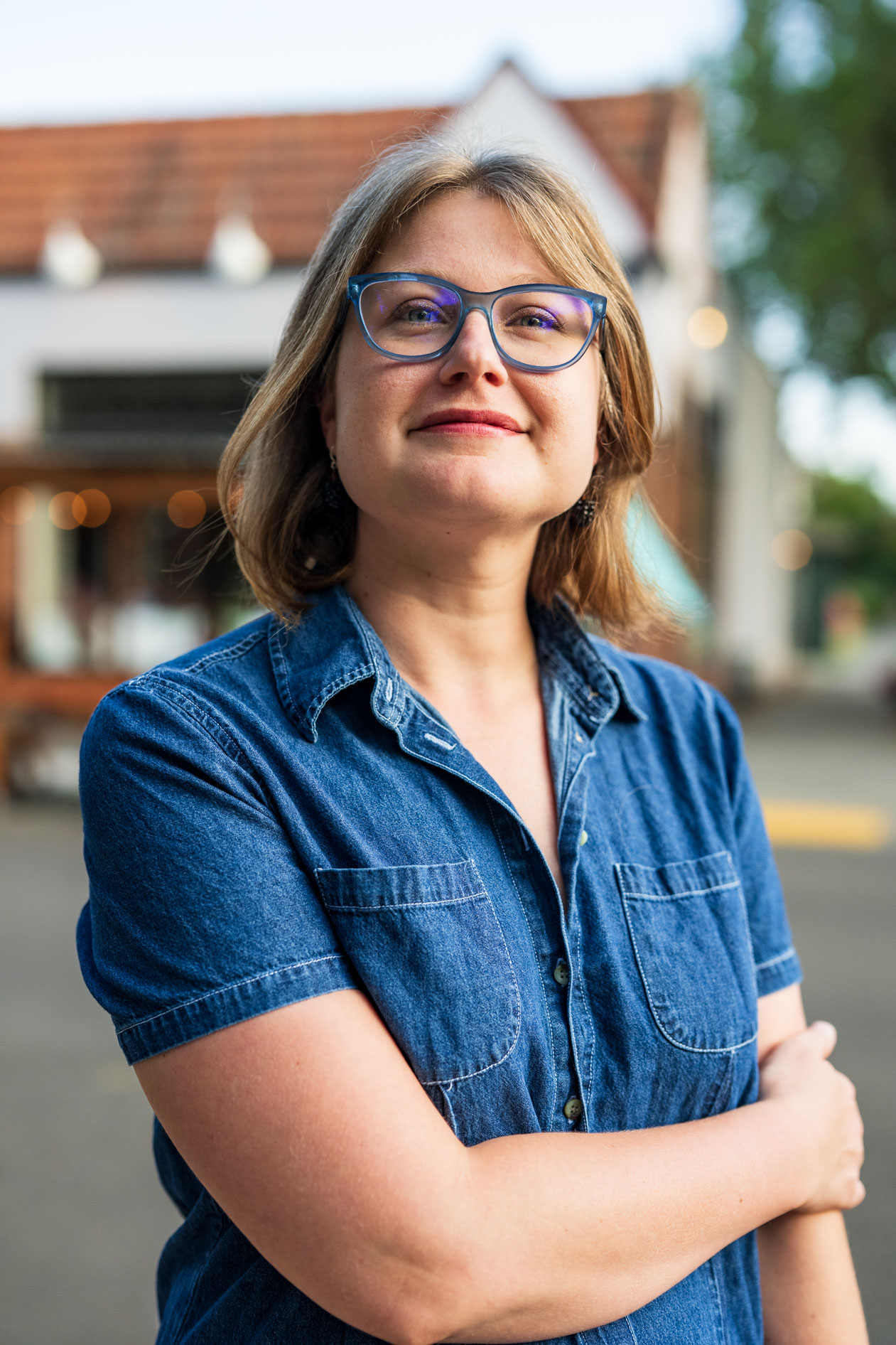 A low-angle outdoor professional headshot of Laura Nash in Portland, Oregon. She is wearing blue glasses and a short-sleeved denim button-down shirt, looking slightly past the camera with a confident expression against a blurred urban background.