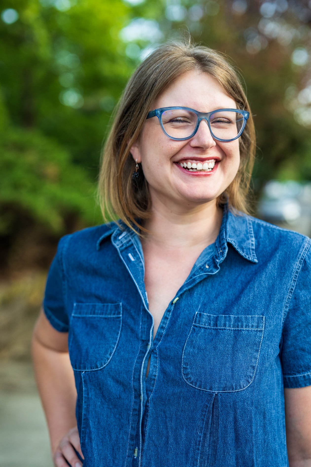 A candid-style professional headshot of Laura Nash laughing. She is wearing blue-framed glasses and a denim shirt, captured in a bright, outdoor setting with vibrant green foliage blurred in the background.