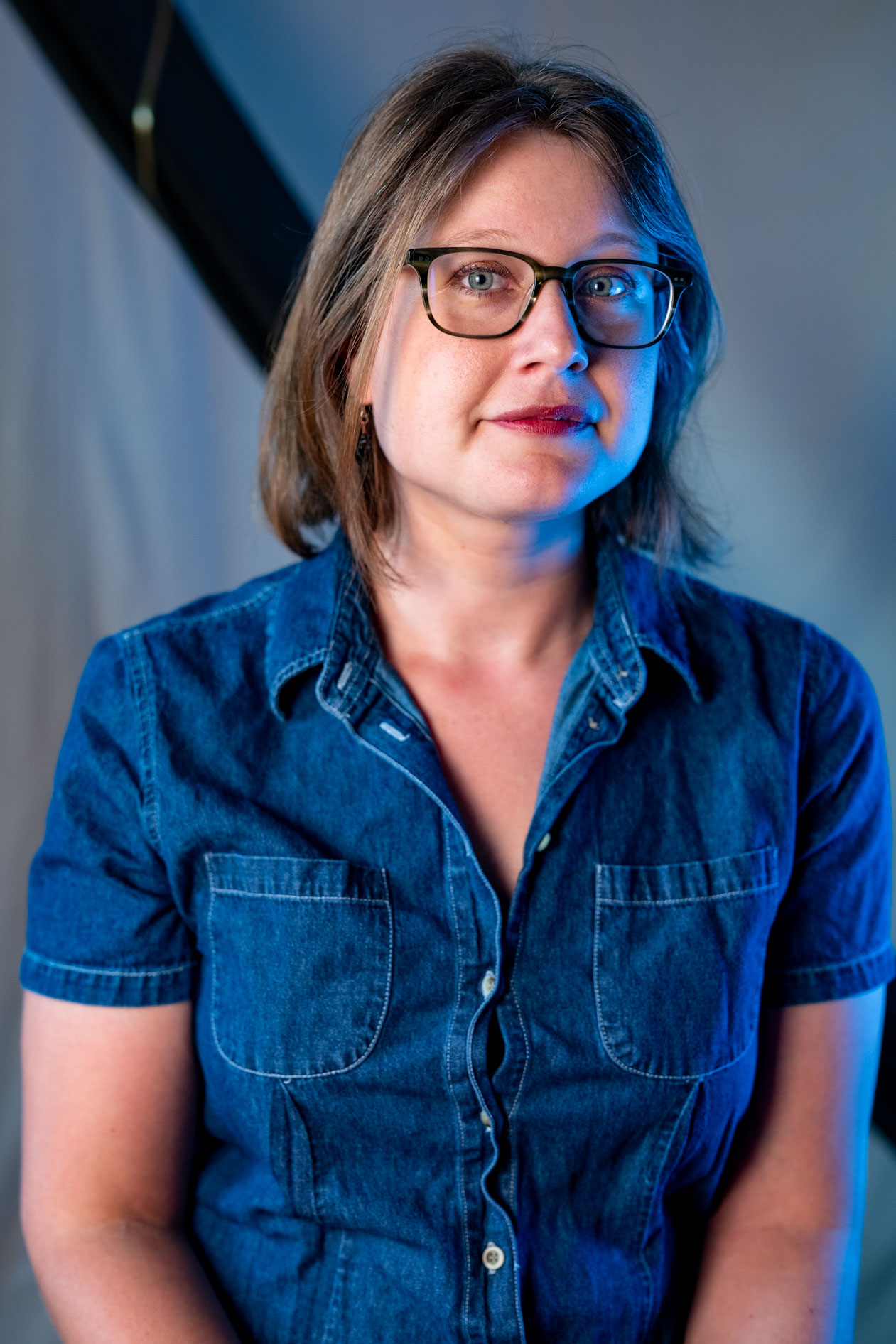 Professional studio headshot of Laura Nash in Portland, Oregon, wearing tortoiseshell glasses and a denim shirt. The portrait features modern, dramatic blue rim lighting against a neutral grey backdrop.