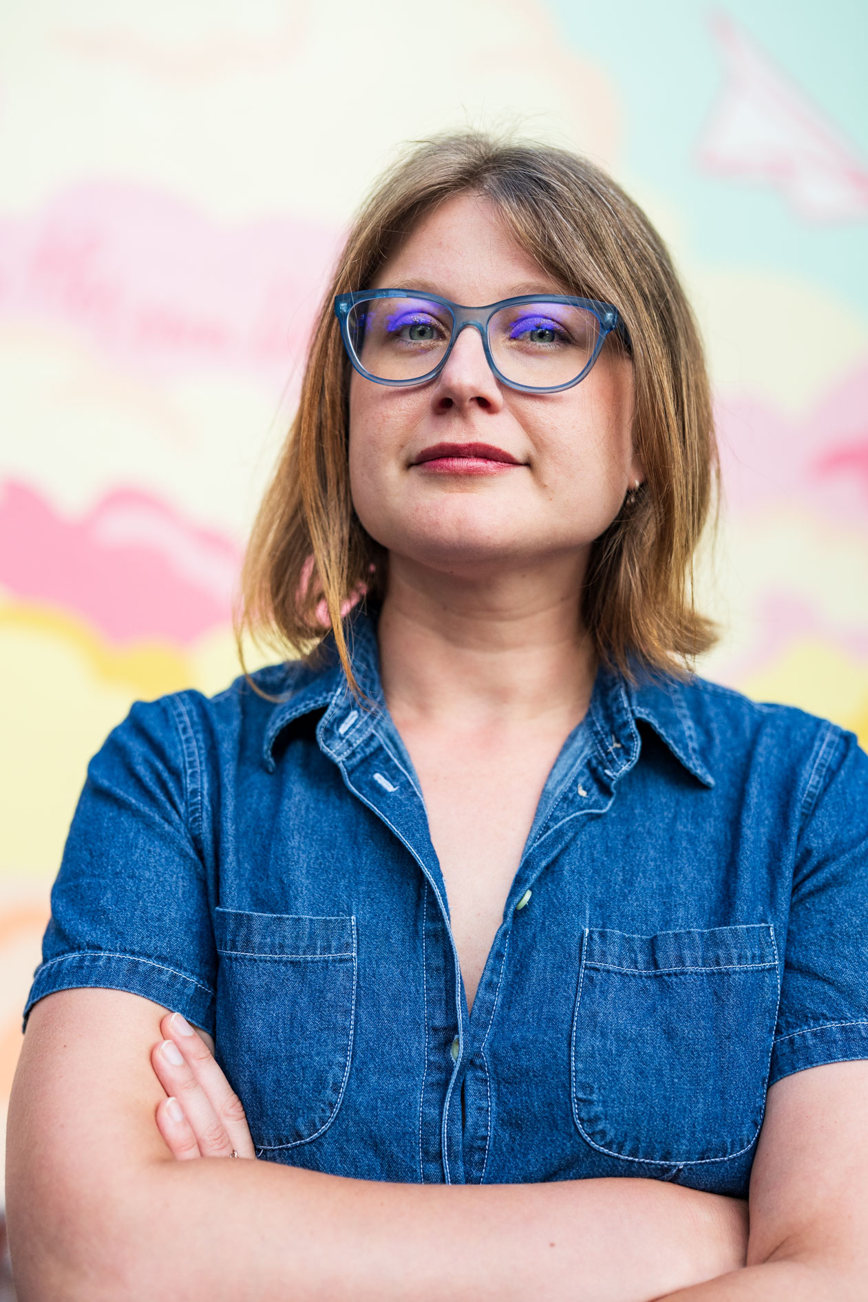 Professional headshot of Laura Nash in Portland, Oregon, wearing blue glasses and a denim shirt. She stands with arms crossed against a colorful, soft-focus mural background featuring shades of pink, yellow, and blue.