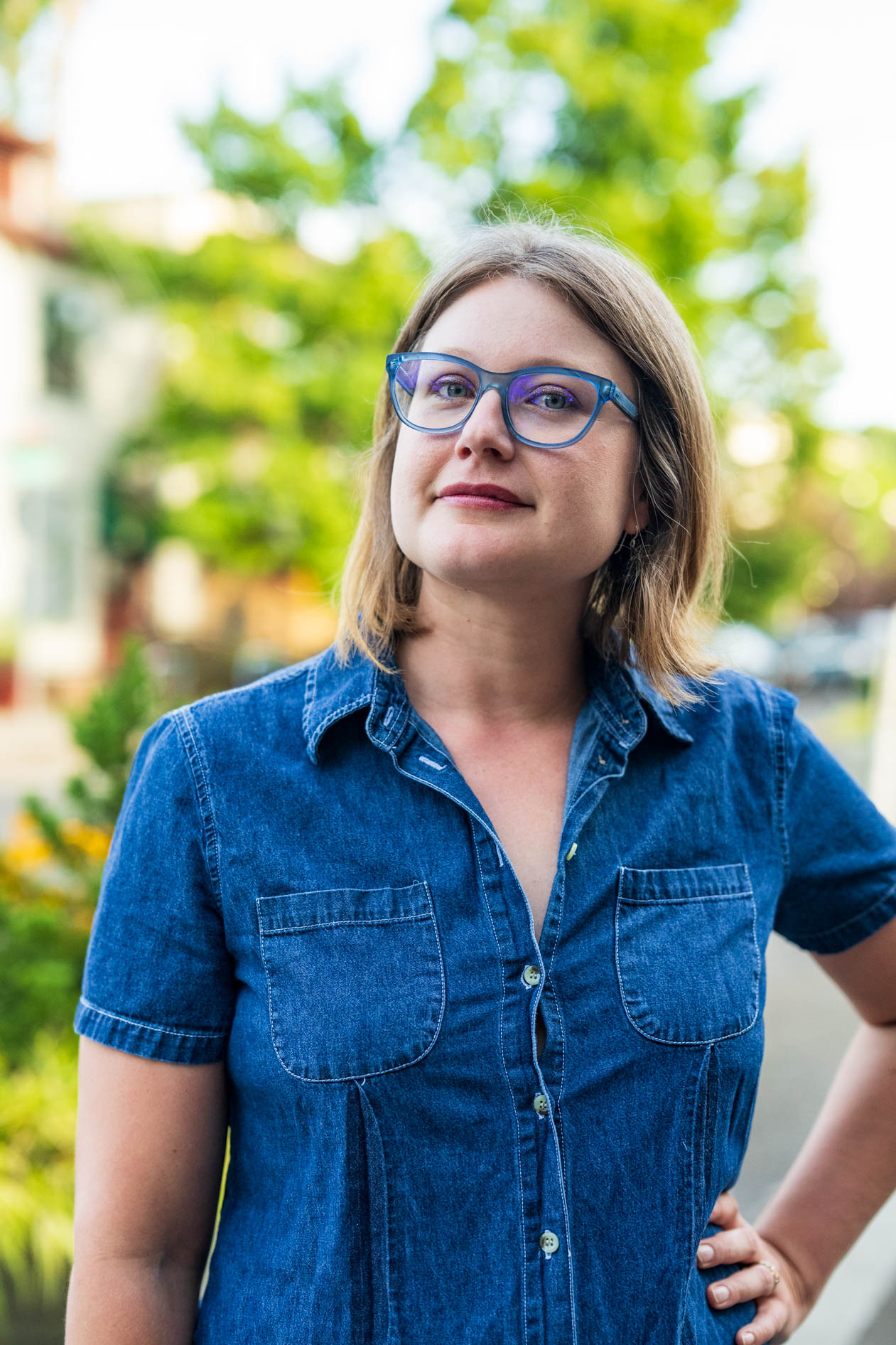 Professional outdoor headshot of Laura Nash in Portland, Oregon. She is wearing blue-framed glasses and a denim button-down shirt, looking directly at the camera with a confident expression against a blurred urban street background with green trees.