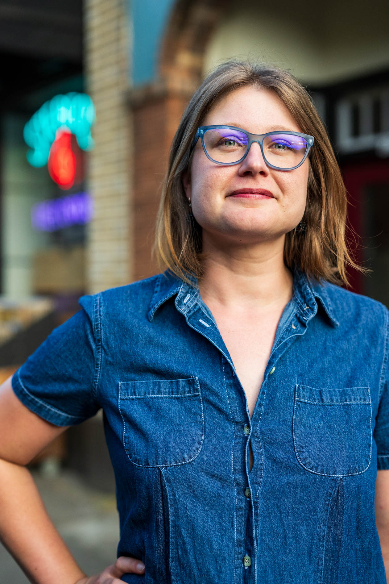 Professional outdoor headshot of Laura Nash in Portland, Oregon, wearing blue glasses and a denim shirt. She poses confidently with a slight smile against a blurred urban background featuring a glowing red and blue neon sign.