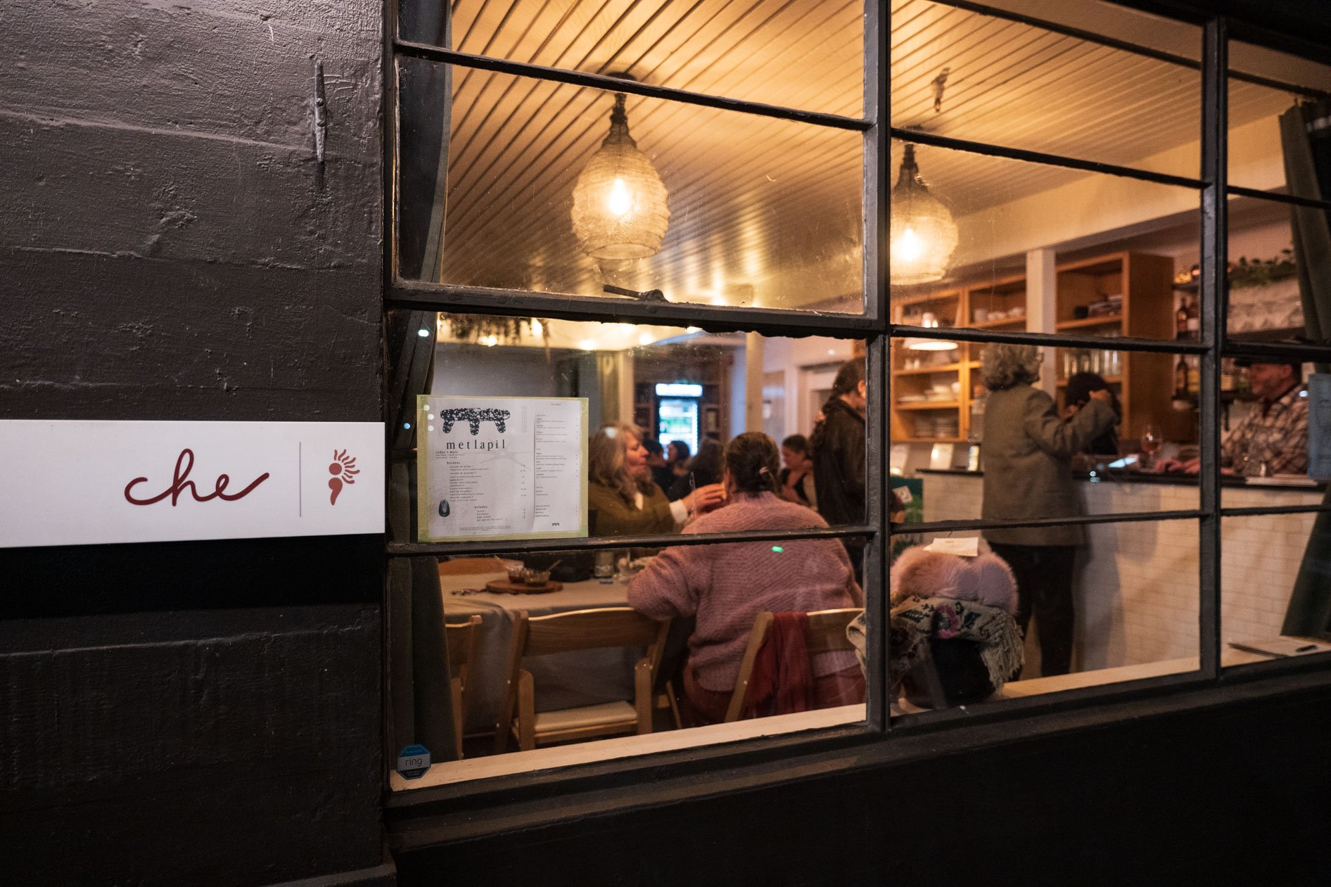 An evening exterior view of Che event space in Portland, Oregon. The warm, lit interior is visible through a large multi-paned window, showing guests gathered at tables for a community Soup Night fundraiser.