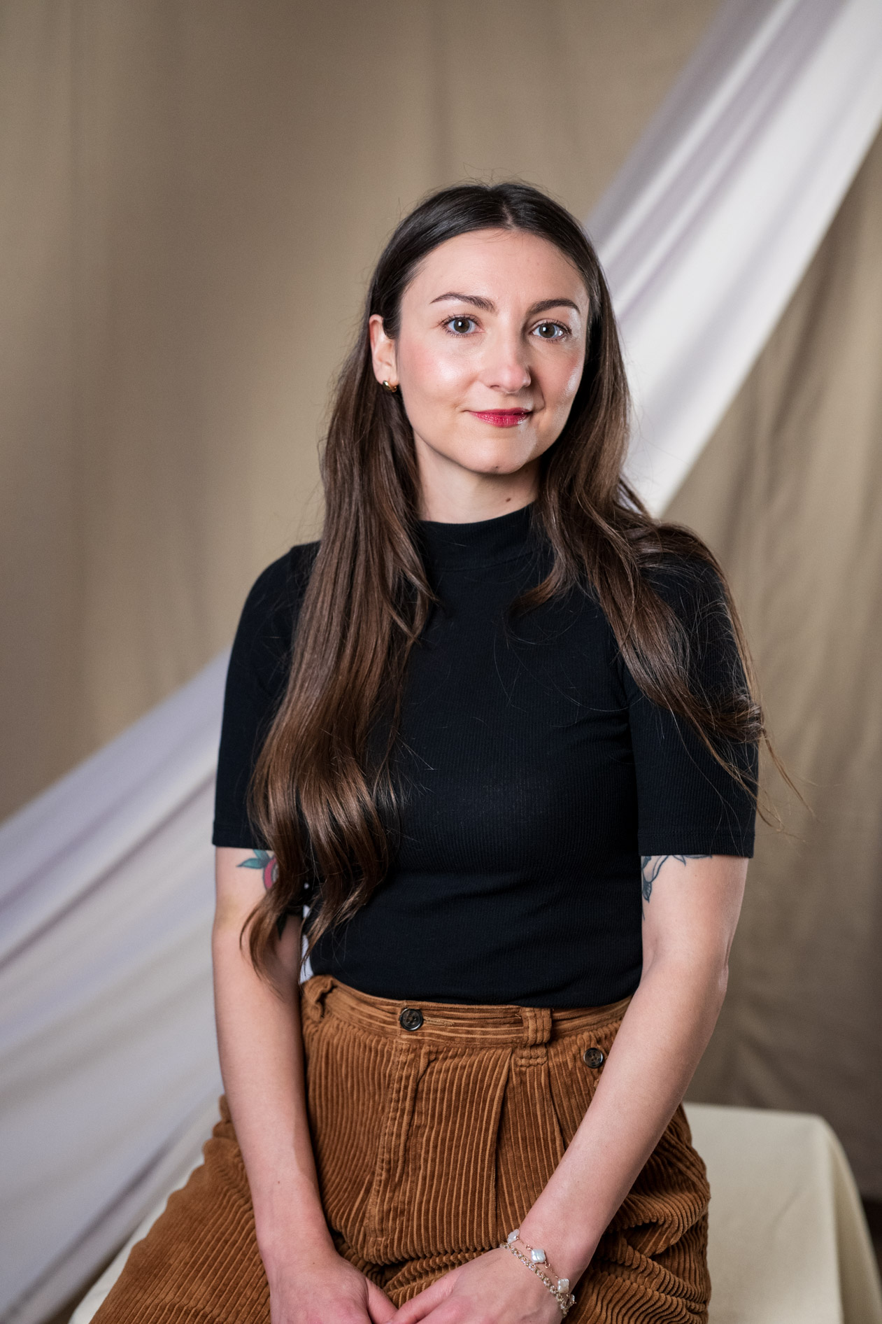 A studio portrait of Miranda Rabuck smiling slightly while seated, wearing a black t-shirt and brown corduroy trousers against a backdrop of draped neutral fabric.