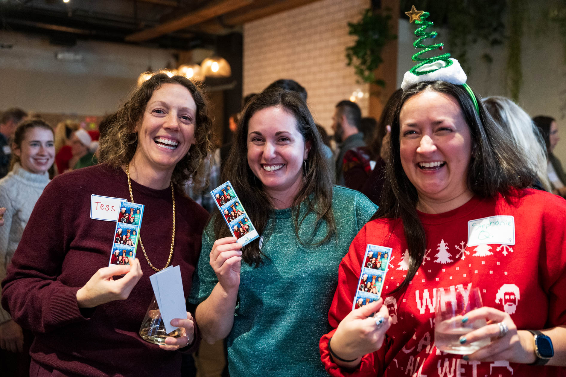 Three people smiling and laughing at a holiday party, holding up vertical photo booth strips and drinks. One person on the right wears a bright red festive sweater and a headband with a small tinsel Christmas tree.