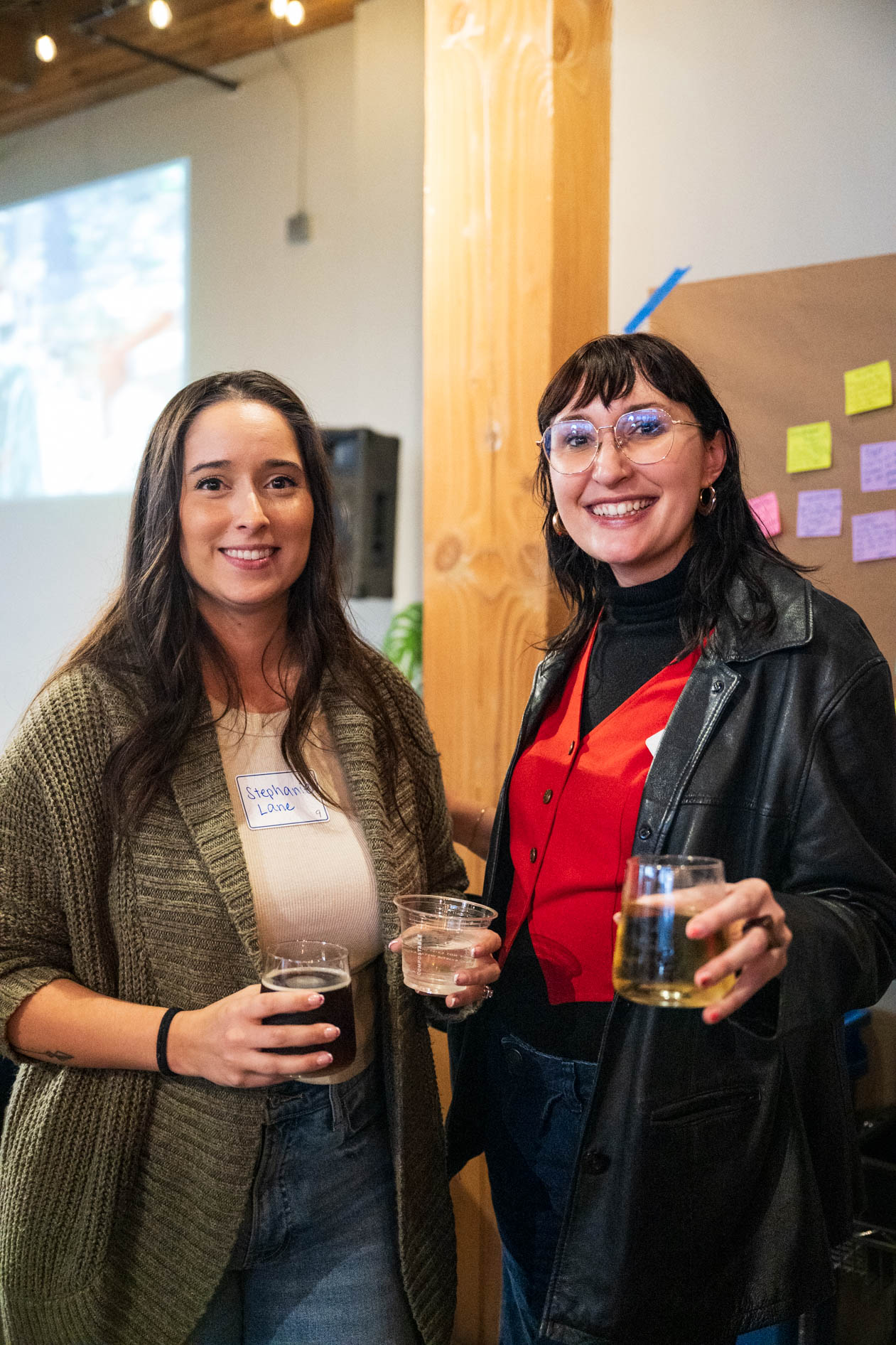 Two people smiling while holding drinks at a holiday party; one wears a long olive green cardigan and the other wears a red vest under a black leather jacket, standing in front of a wooden pillar with sticky notes in the background.