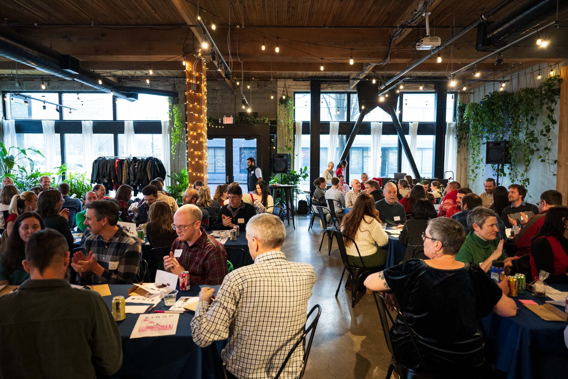 A large group of people seated at blue tables in a bright, industrial-style event space with string lights and high ceilings during a holiday party.
