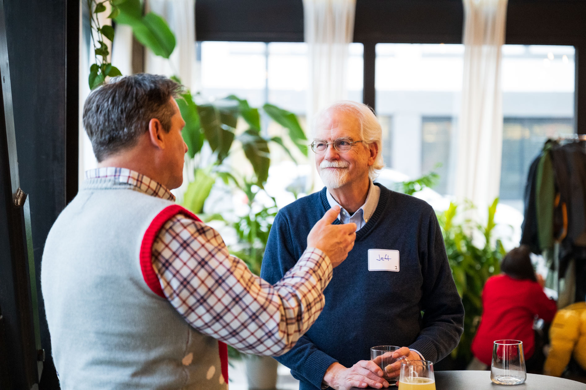 Two men in festive sweaters talking and holding drinks during a holiday party in front of a window with indoor plants.