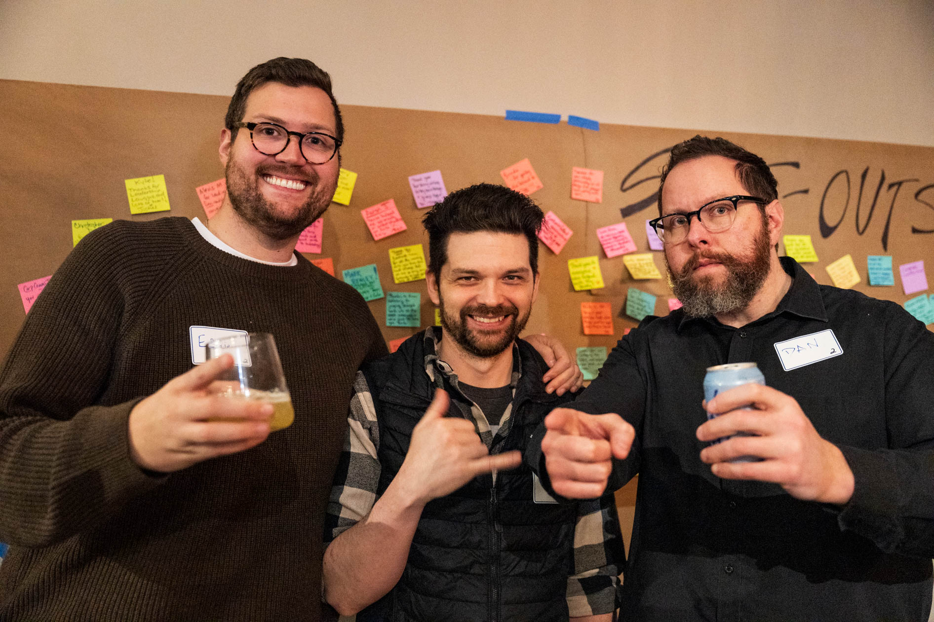 Three men smiling and posing with drinks in front of a "Shout-outs" wall covered in colorful sticky notes at a holiday party in Portland.