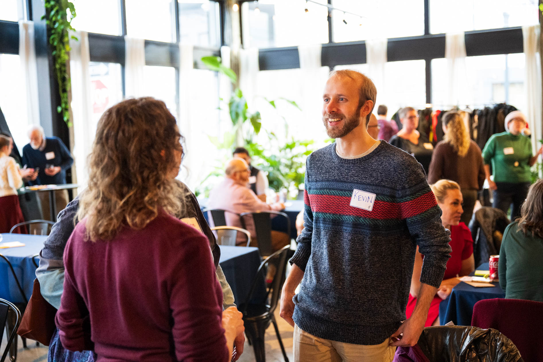 A person named Kevin in a striped holiday sweater smiles while talking to another person in a purple top at a party in a bright Portland event space.
