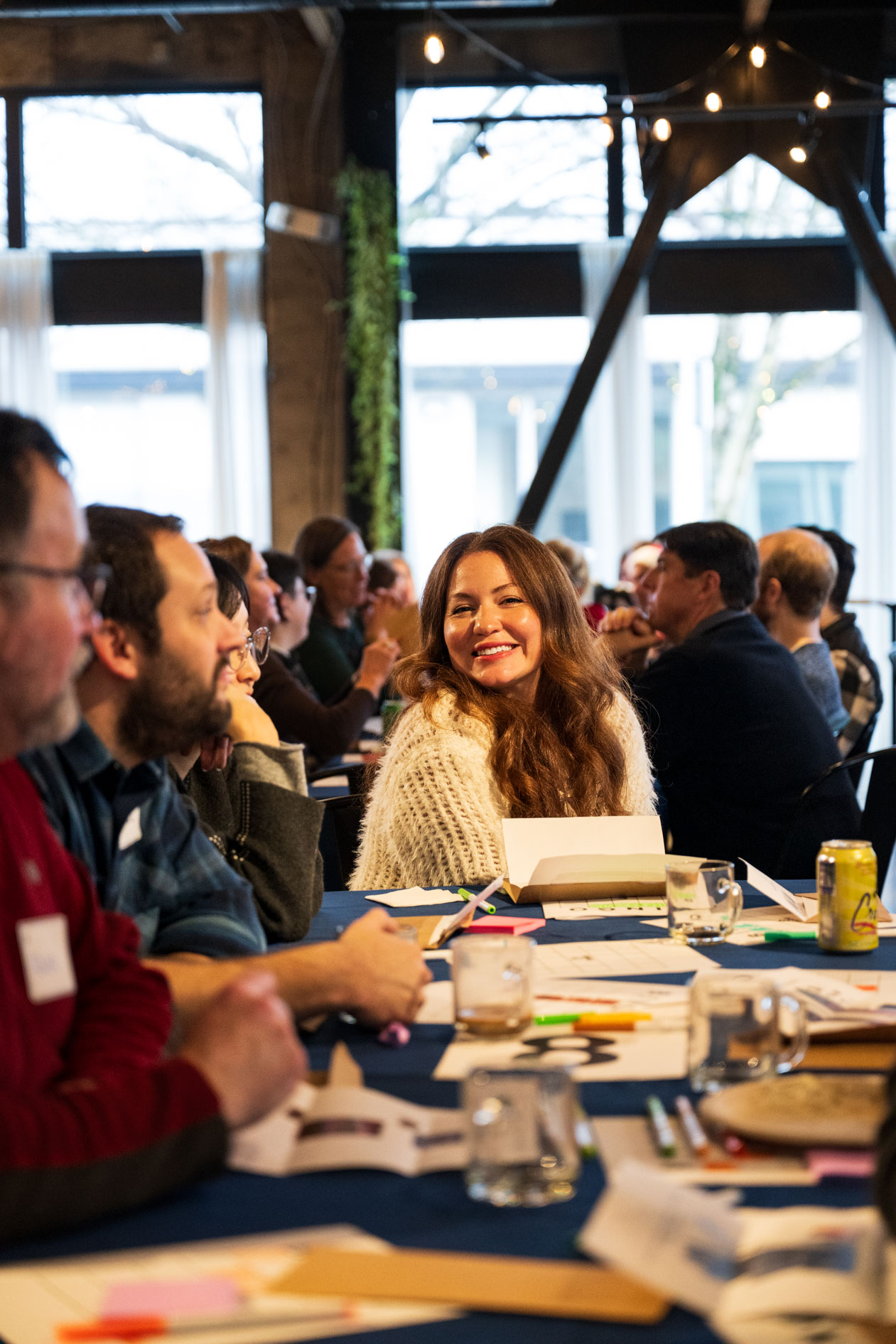 A person in a white textured sweater smiles while seated at a blue table during a holiday party in a bright Portland event space.