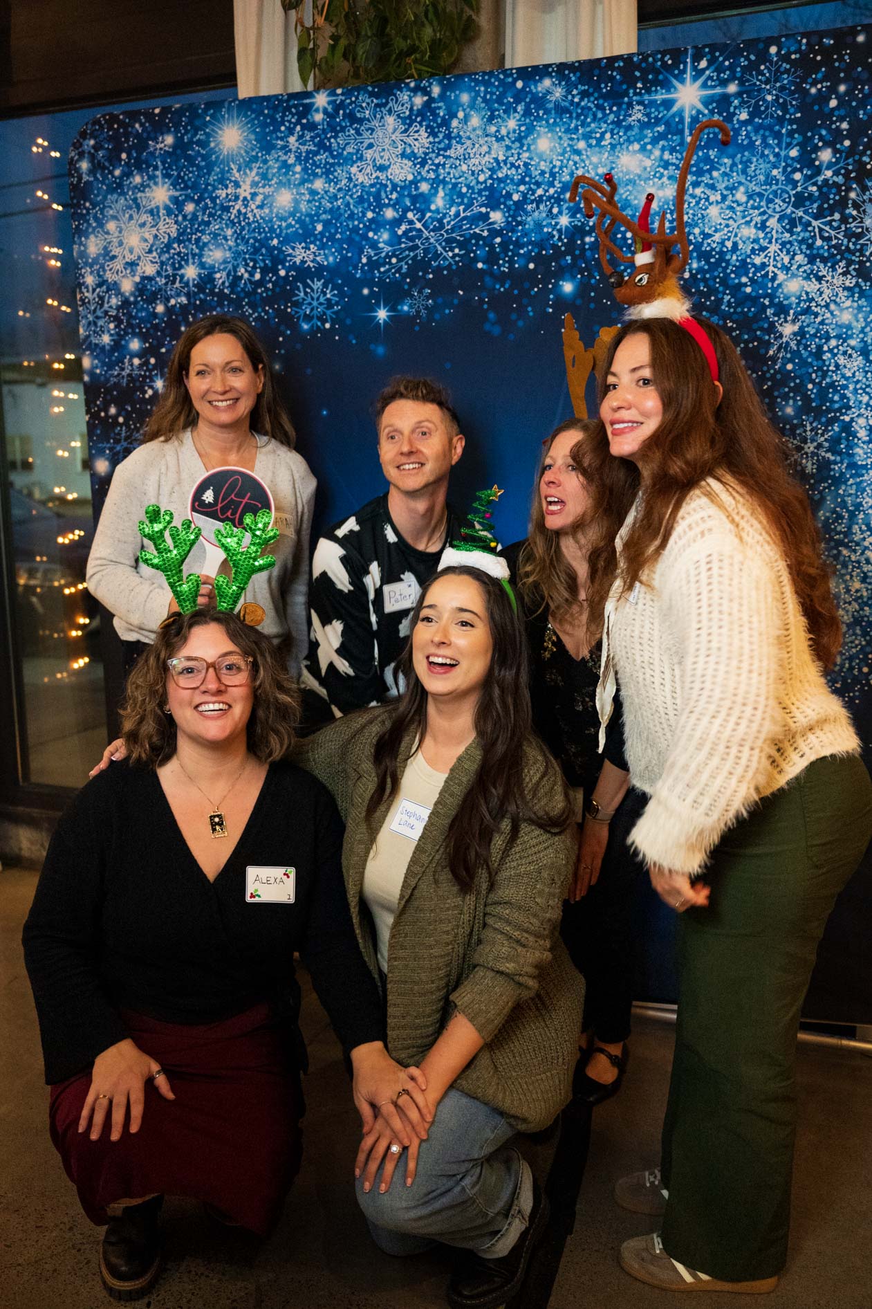A group of people posing and laughing in front of a blue snowy backdrop at a holiday party in Portland; several people wear festive headbands, including reindeer antlers and tinsel trees.