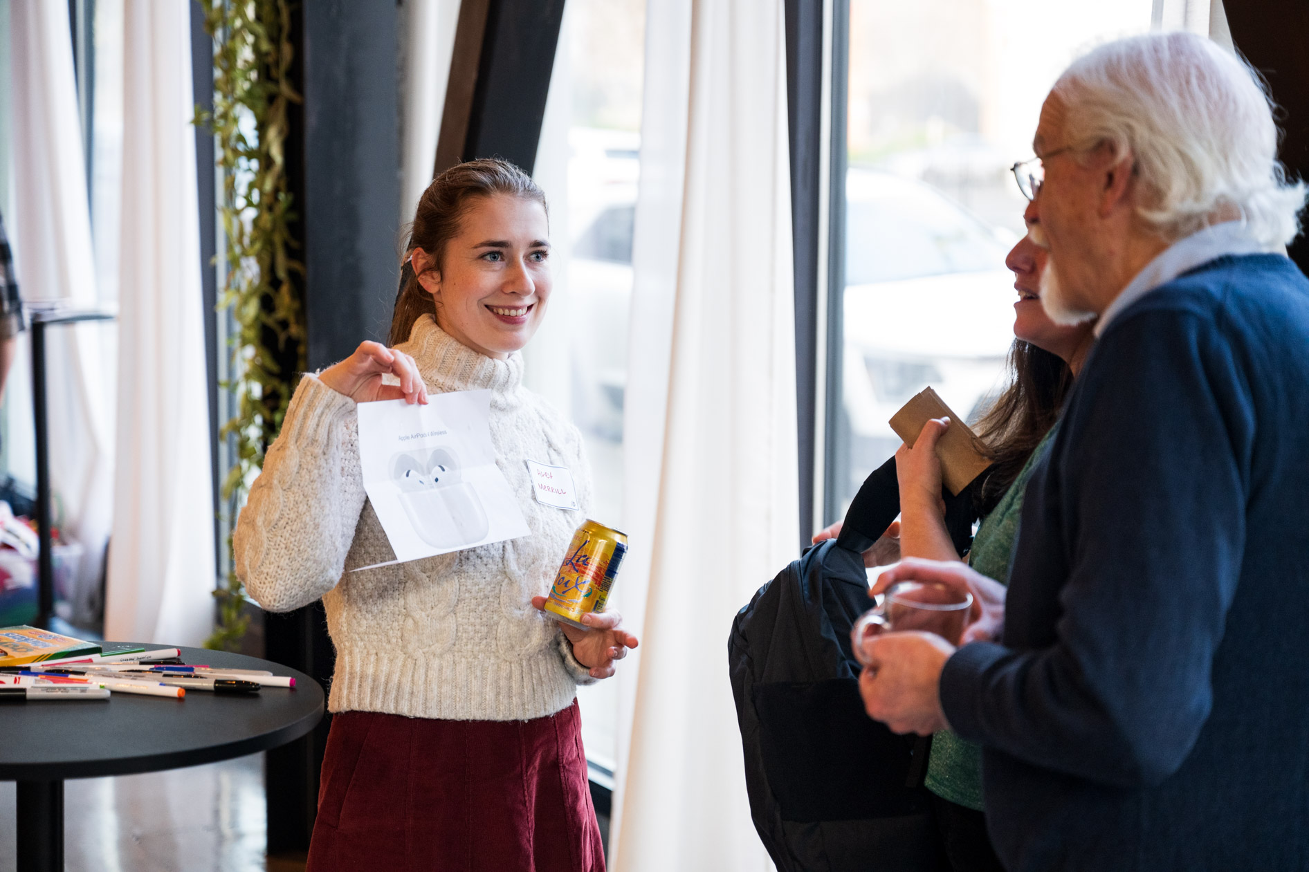 A person in a cream-colored cable-knit sweater holding up a paper showing an AirPods prize while talking to guests at a holiday party in Portland.