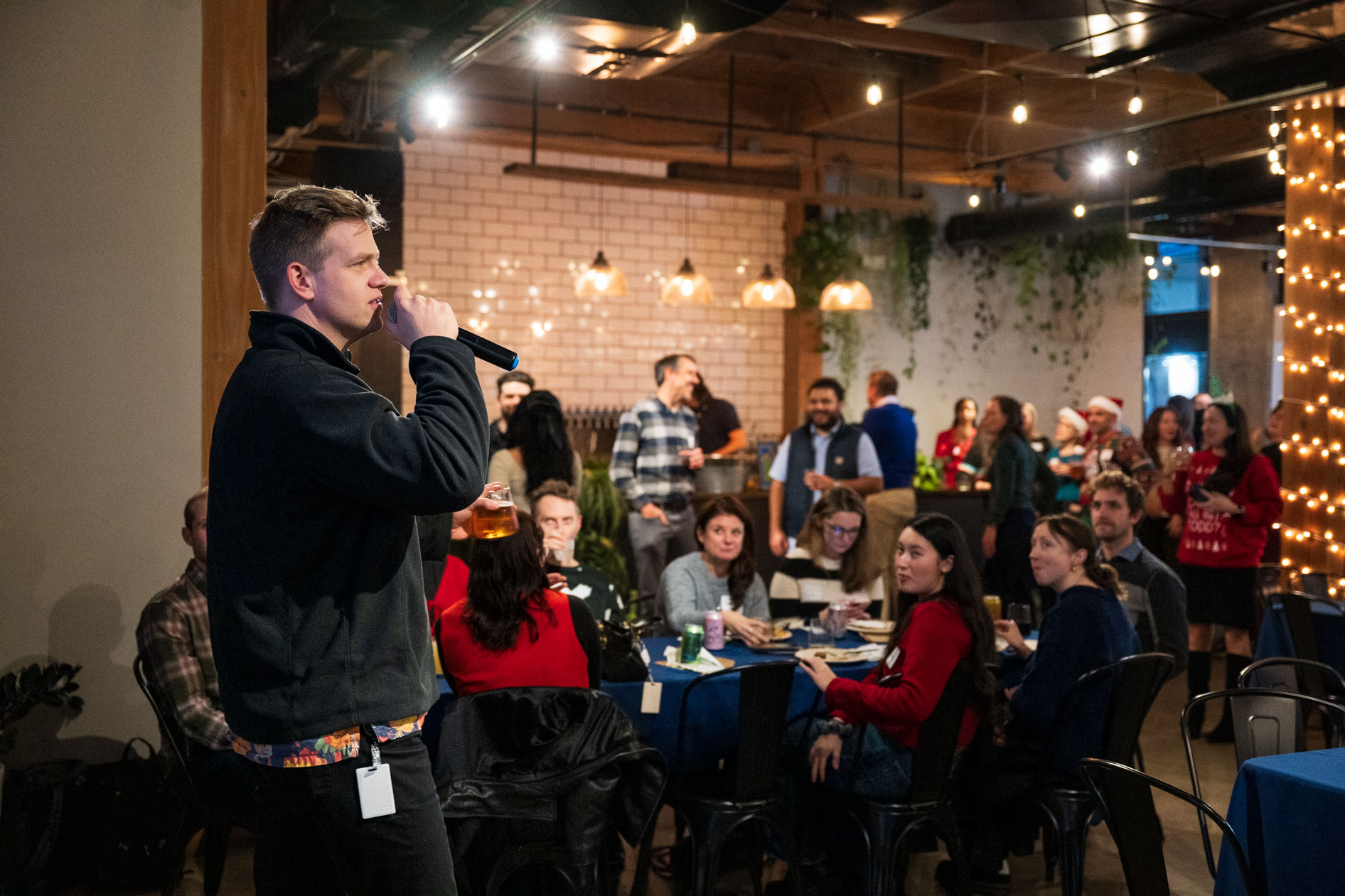 A person speaking into a microphone while holding a drink, addressing a room of guests seated at blue tables during a holiday party in a Portland event space.