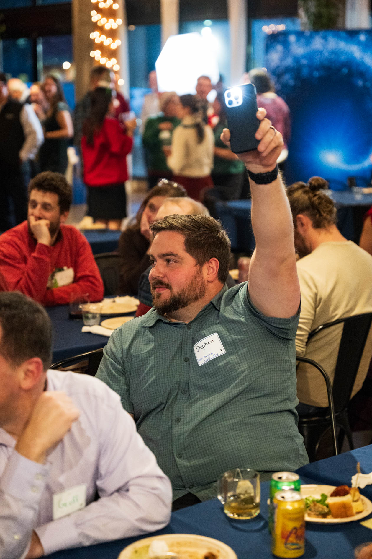 A person named Stephen seated at a table raises a smartphone with the flashlight turned on during a holiday party in a Portland event space.