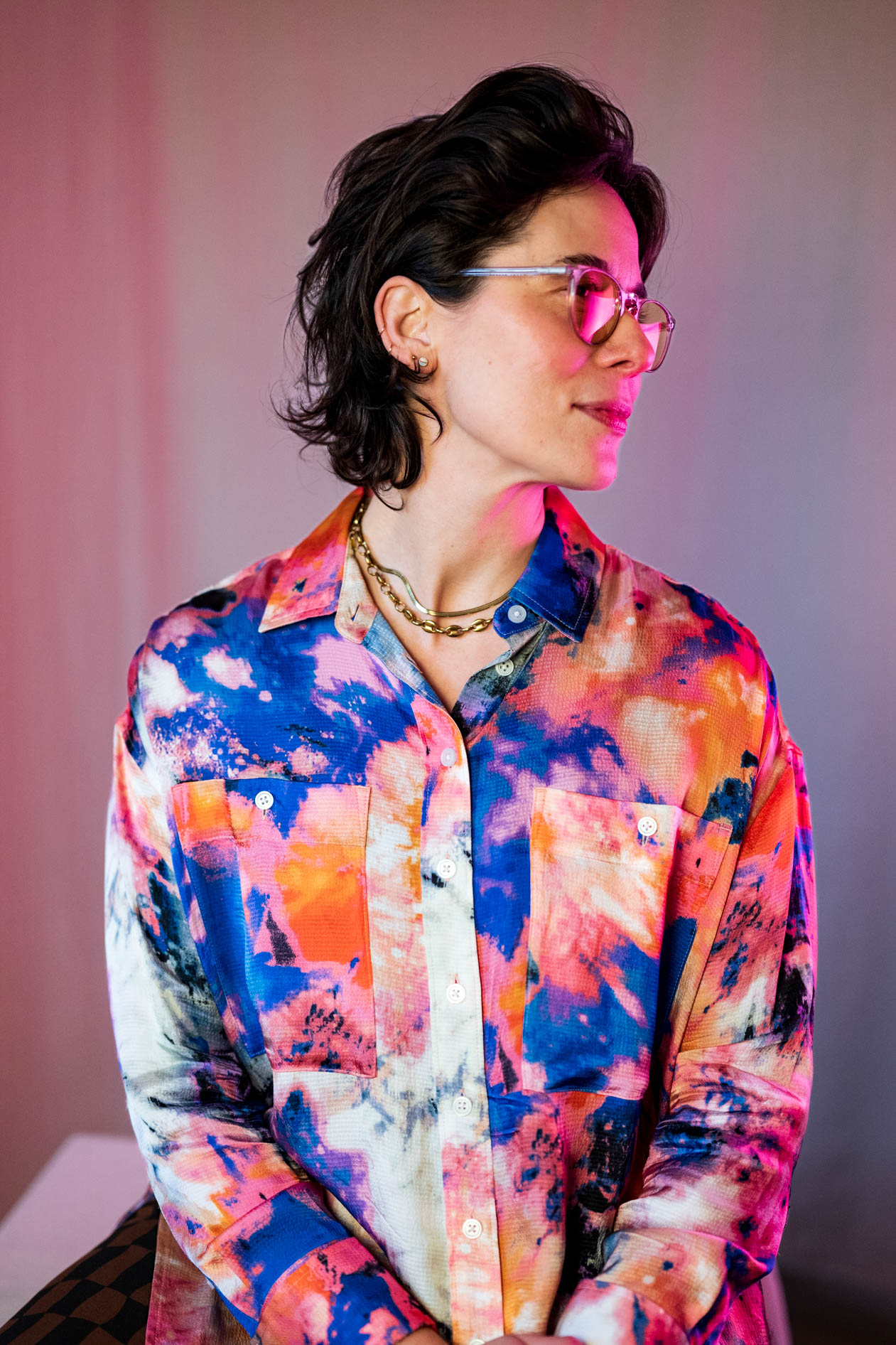 A person in a colorful tie-dye shirt and yellow-tinted glasses looking to the side under pink and blue studio lighting.