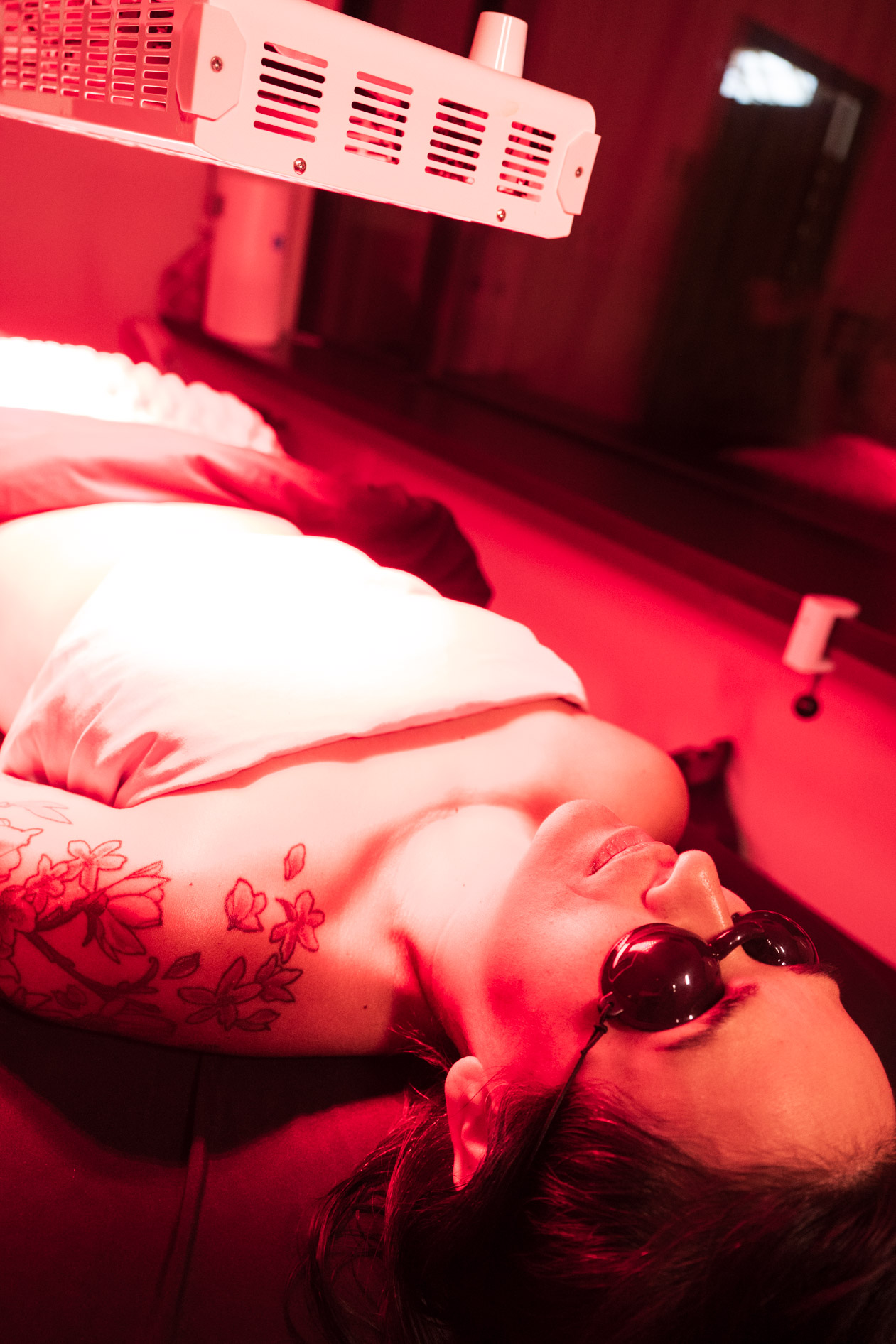 A person with a floral shoulder tattoo wearing protective goggles lies on a treatment table under a red LED light therapy panel in a beauty studio in NE Portland.