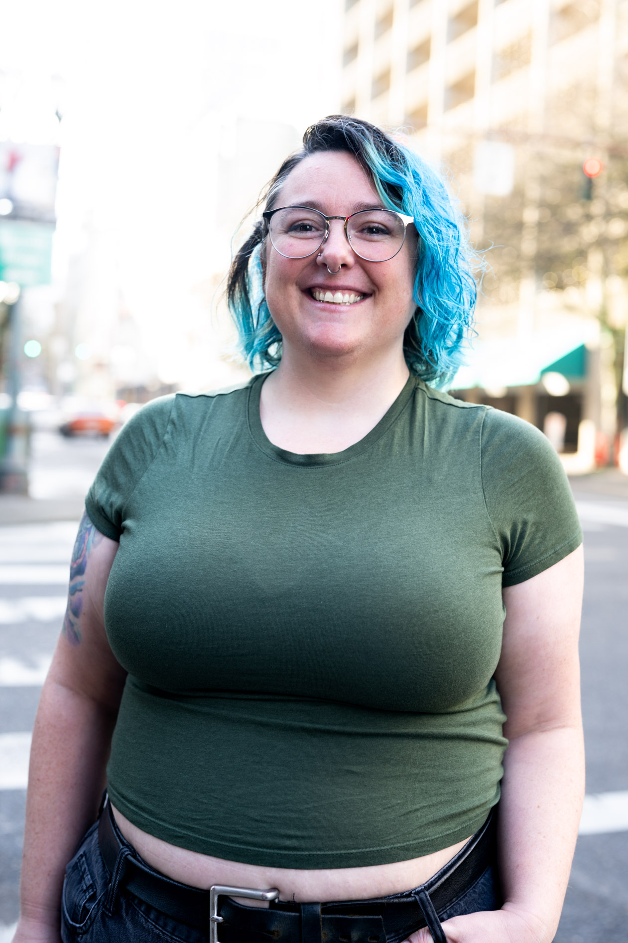 A professional portrait of a person with bright blue hair and glasses, wearing a dark green t-shirt and smiling while standing on an urban street in Portland.
