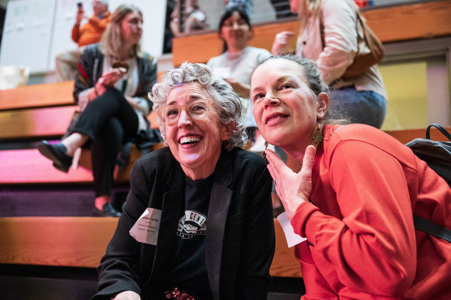 A candid close-up of two people smiling and looking toward a presentation at the B Bash event. One attendee wears a Grand Central Bakery t-shirt and blazer, reflecting the community of values-led business leaders at Wieden + Kennedy.