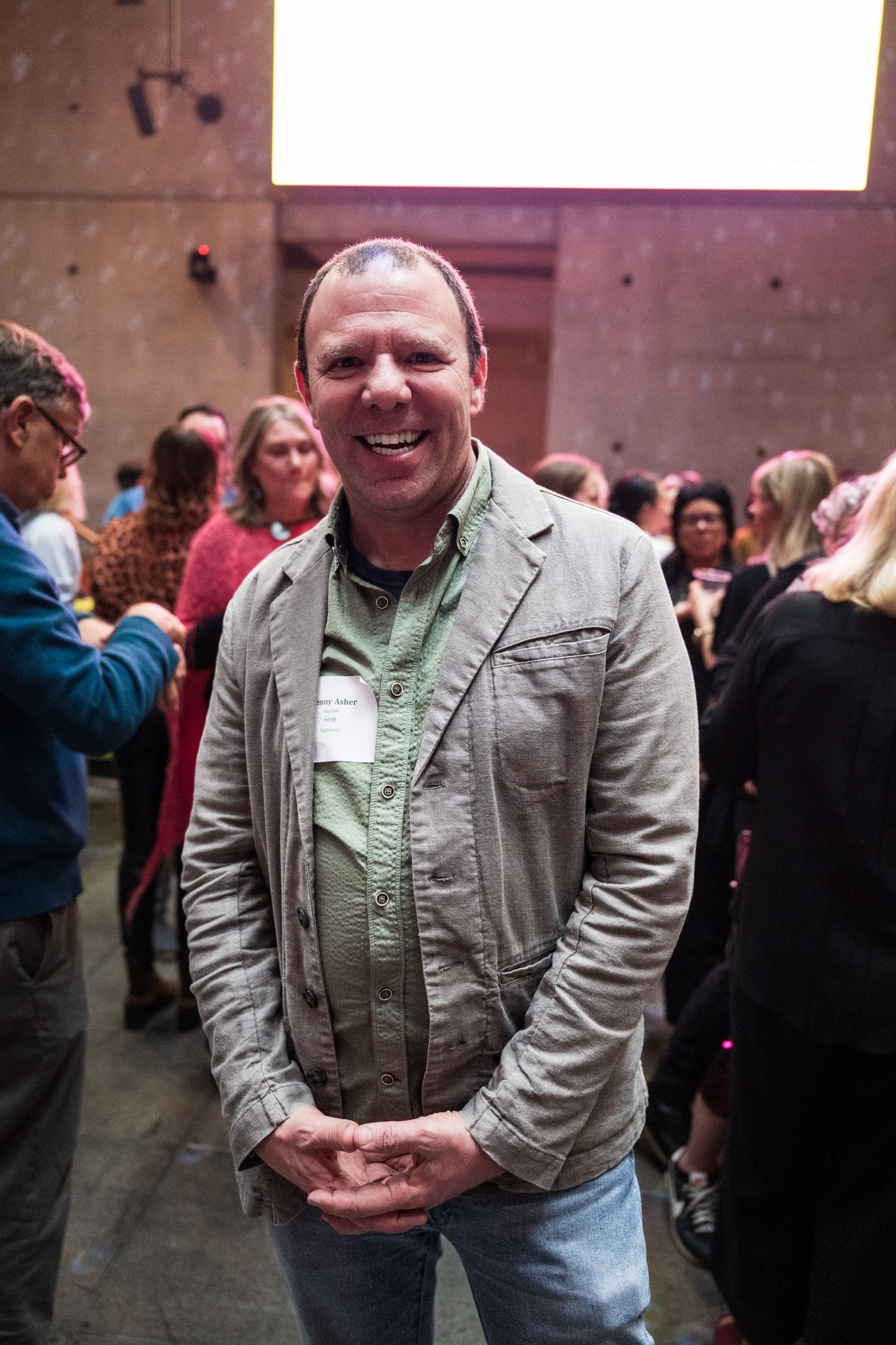 A portrait of a smiling attendee in a grey blazer and green button-down shirt at the B Bash event. He stands in the Wieden + Kennedy atrium during the On Purpose Oregon launch celebration.