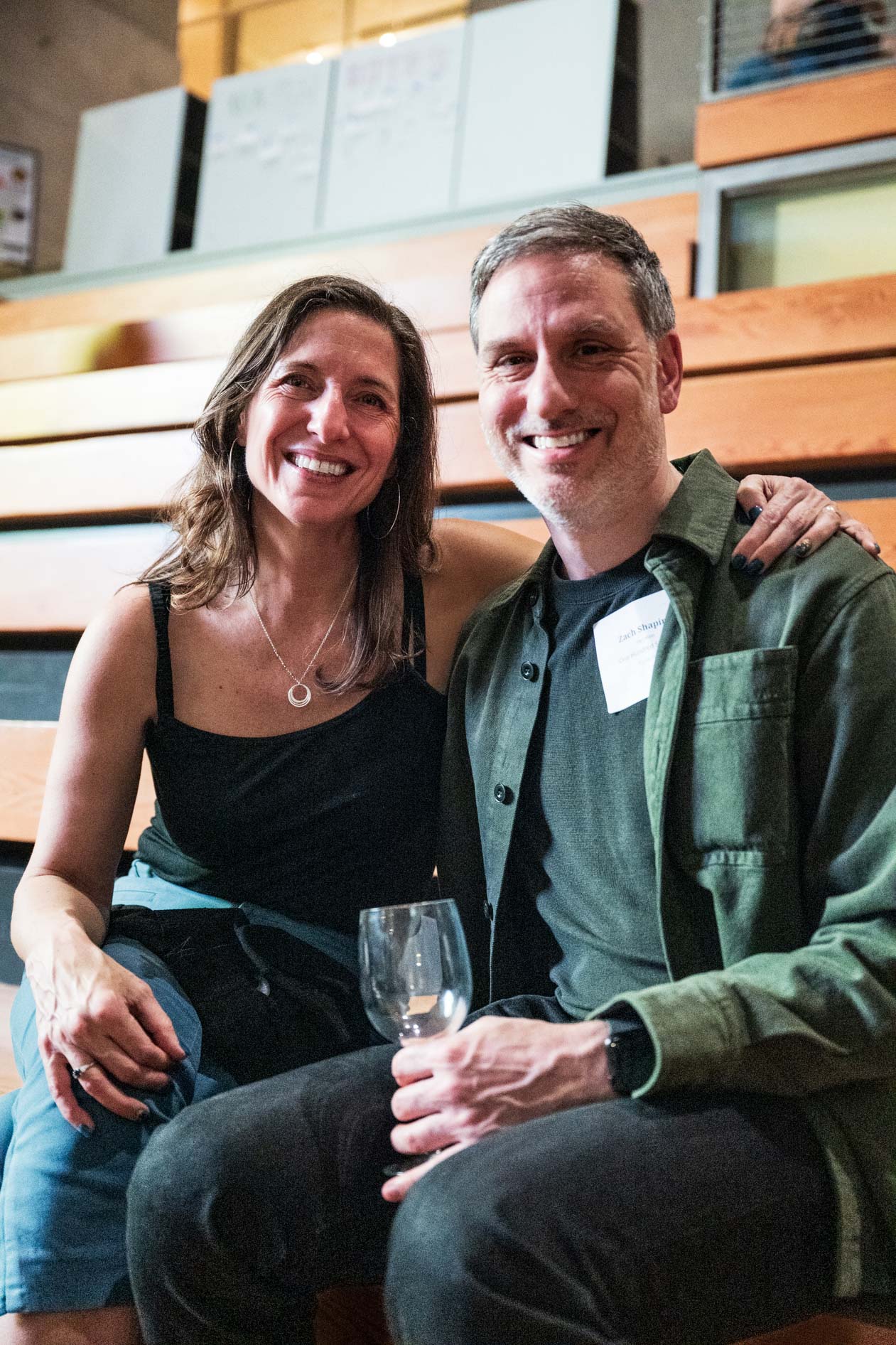 Two attendees seated on wooden bleachers at the B Bash event. One wears a black tank top and silver necklace, and the other wears an olive green shirt jacket, capturing a friendly moment at Wieden + Kennedy during the On Purpose Oregon launch.