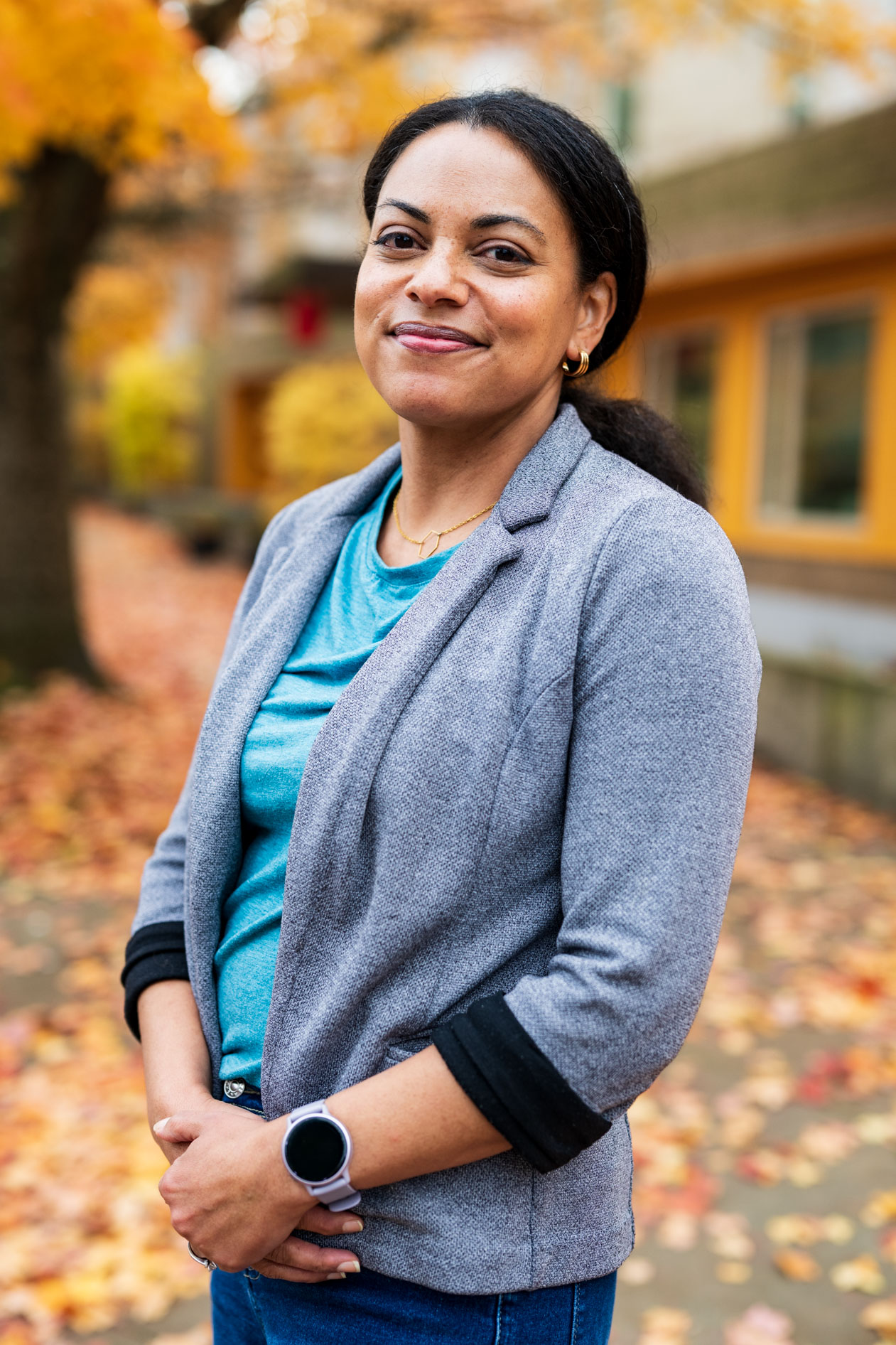 A professional headshot of a person smiling calmly outdoors during autumn. They are wearing a grey blazer over a teal top, with gold jewelry and a smartwatch, standing against a blurred background of yellow fall leaves and a building.