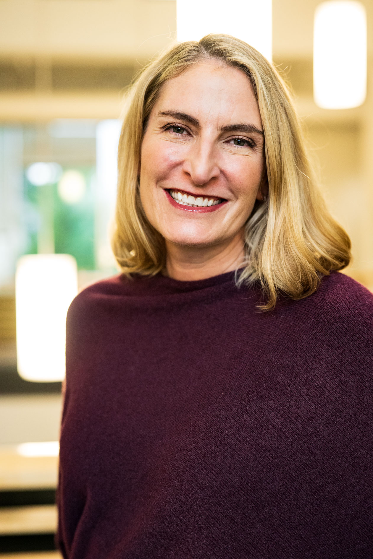A professional headshot of a person with blonde hair smiling warmly. They are wearing a maroon sweater against a softly blurred indoor background with warm lighting, capturing an approachable and friendly presence for a Planned Parenthood case study.