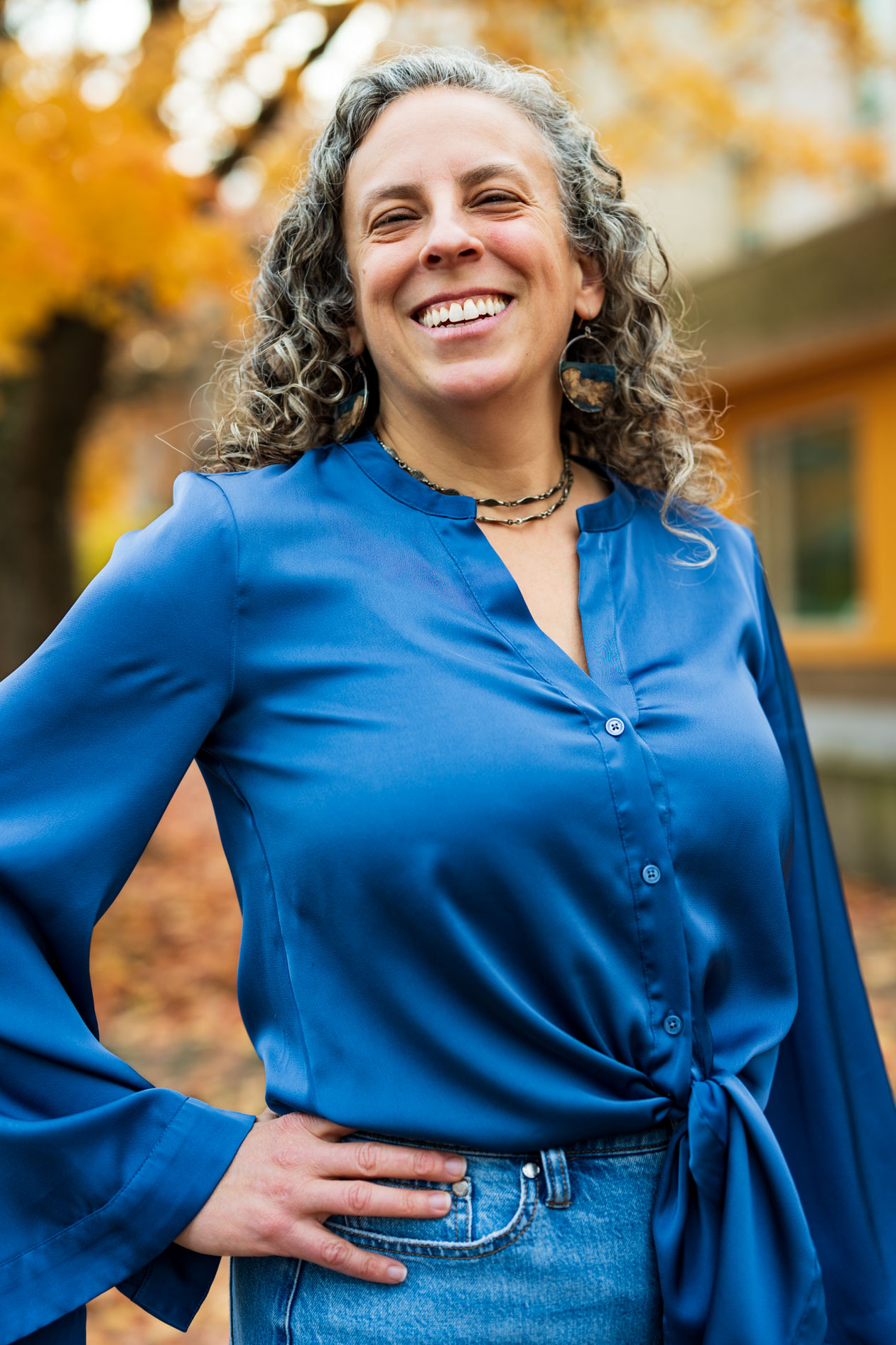 A person with curly hair laughs joyfully during an outdoor professional headshot session. They are wearing a bright blue silk blouse and large hoop earrings, standing with a hand on their hip against a blurred background of autumn leaves and a building.